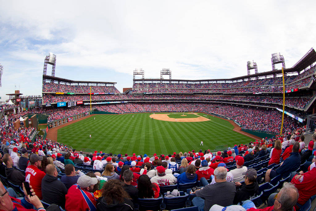 Phillies 桌布 壁紙 — 一個擠滿球迷的棒球場，球迷身穿紅色衣服，在陰天底下歡呼，球員們在綠色場地上比賽。