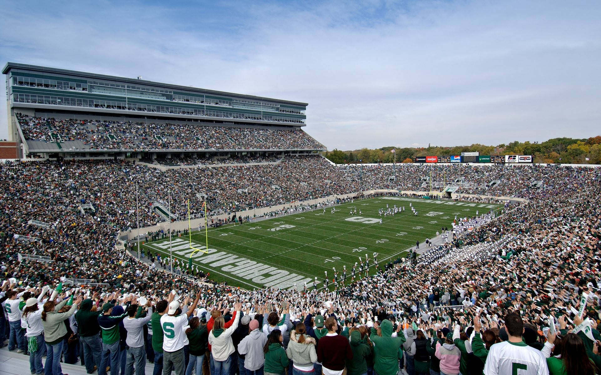 Michigan State Spartans 桌布 壁紙 — 一個擠滿球迷的足球場，球迷身穿綠色和白色衣服，在部分多雲的天空下為場上列隊的球員歡呼。