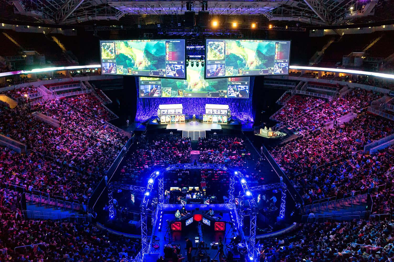 Gaming Tournaments 桌布 壁紙 — 一個擠滿觀眾的場館在紫色和藍色燈光下閃耀，粉絲們正在巨型螢幕上觀看一場大型電競錦標賽。