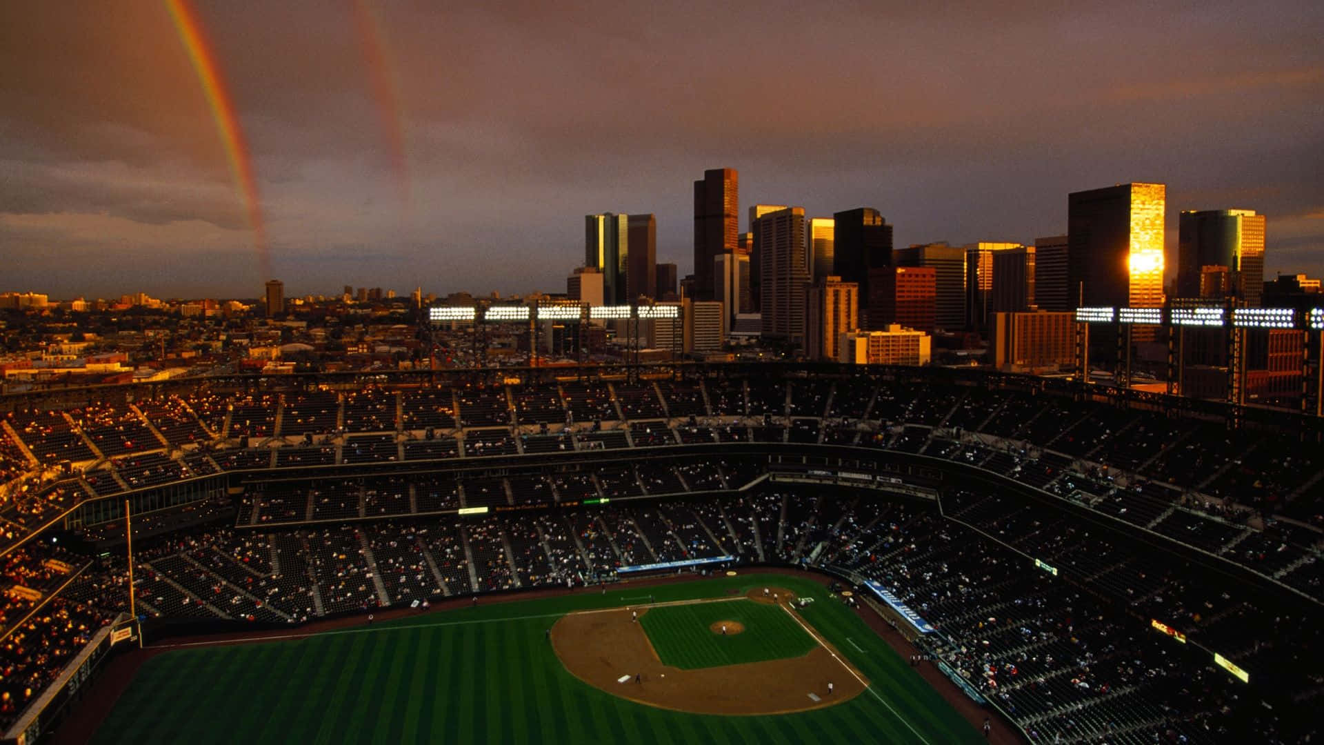 Baseball Stadium 桌布 壁紙 — 一個擠滿觀眾的棒球場，在黃昏的城市天際線上，一道戲劇性的雙彩虹拱起。