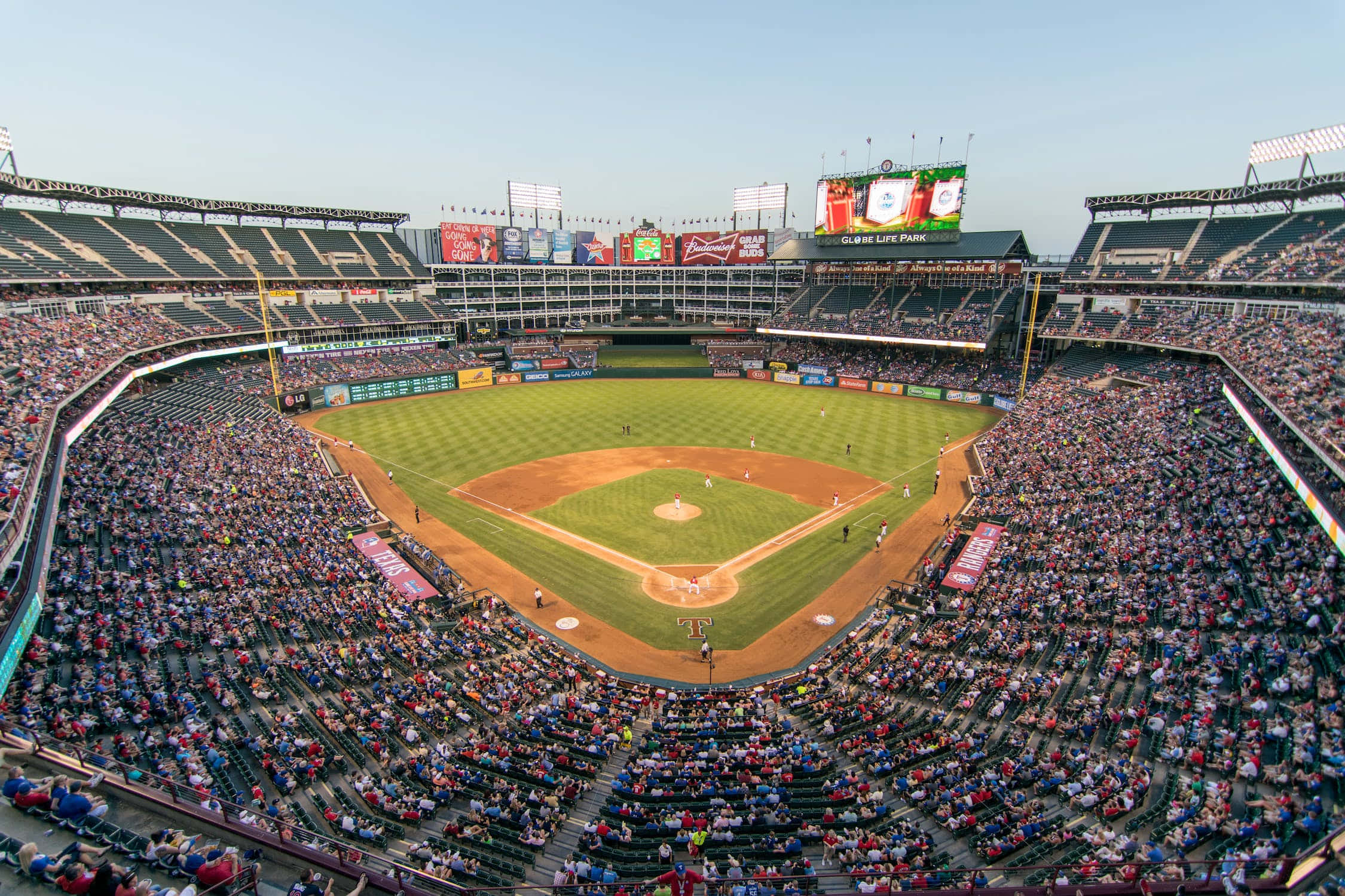 Baseball Field 桌布 壁紙 — 一個擠滿觀眾的棒球場的鳥瞰圖，場地綠草如茵，球迷在晴朗的藍天下歡呼。
