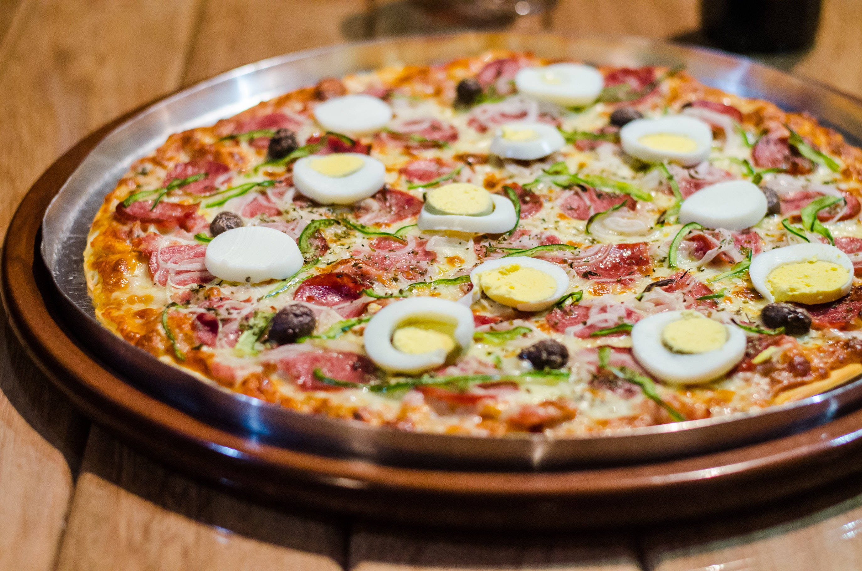 Pizza 桌布 壁紙 — 一個新鮮出爐的披薩，上面鋪有切片水煮蛋、橄欖和火腿，放在木桌上的金屬托盤上。