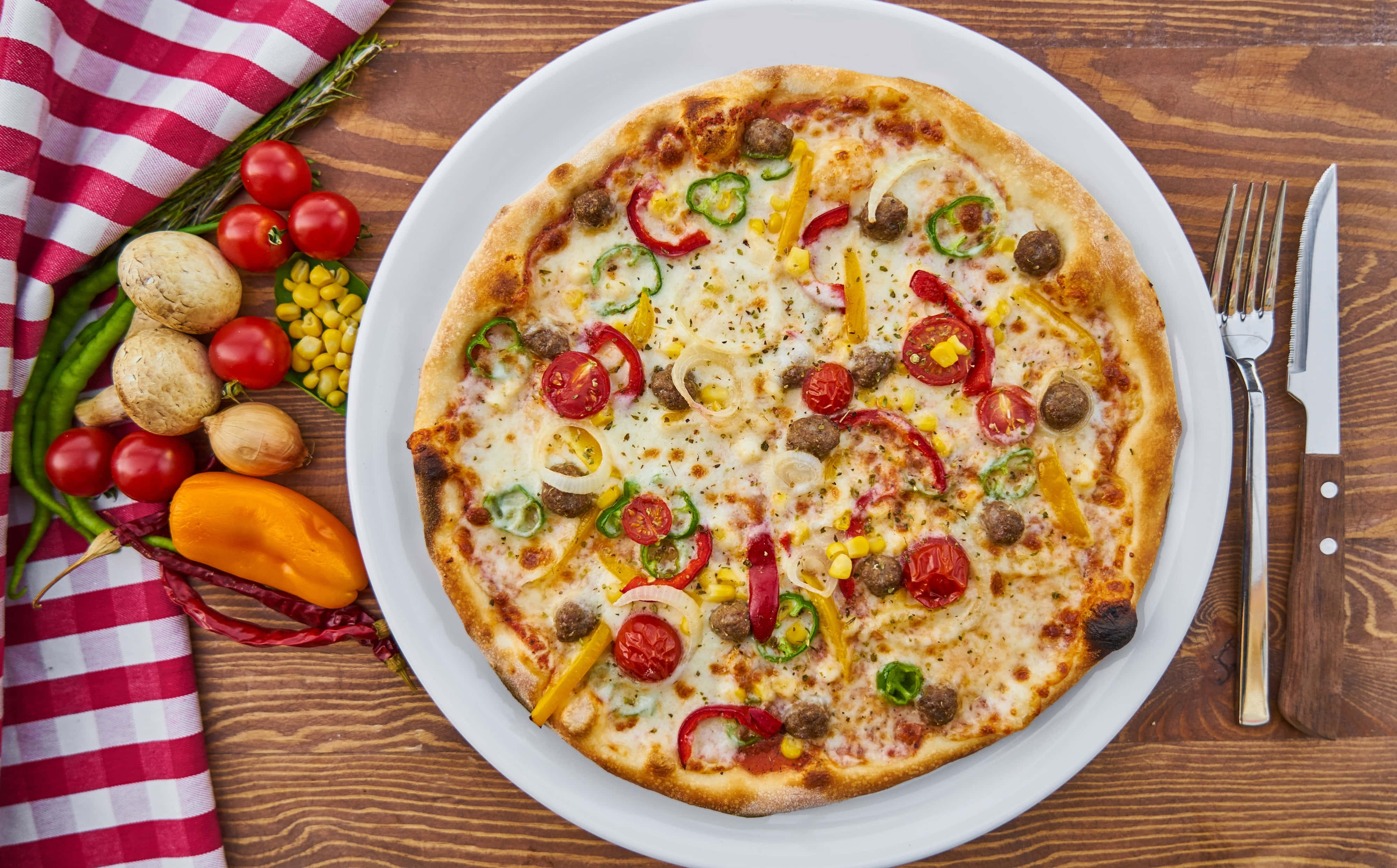 Pizza 桌布 壁紙 — 一個新鮮出爐的披薩，上面鋪有香腸、青椒、玉米和櫻桃番茄，放在白色盤子上，旁邊有紅色格子餐巾和新鮮食材，置於木桌上。