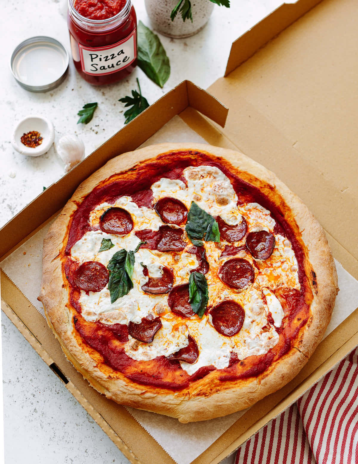 Pizza 桌布 壁紙 — 一個新鮮出爐的義大利辣味香腸披薩，上面有融化的起司和羅勒葉，放在一個打開的紙板箱裡，旁邊有一罐披薩醬和蒜瓣。