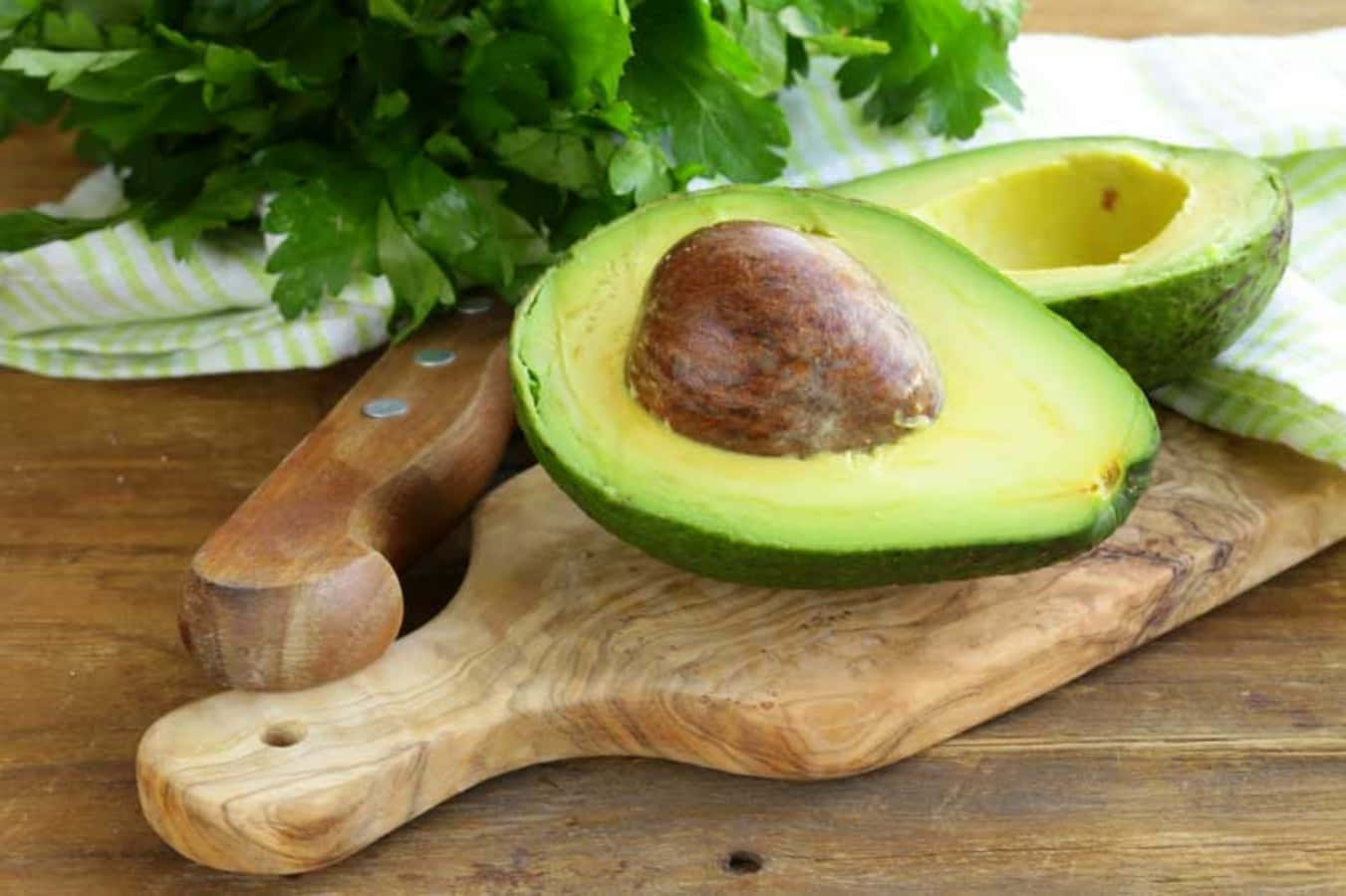 Avocado 桌布 壁紙 — 一個新鮮切開的酪梨，帶有一個大的棕色果核，放在木砧板上，旁邊有一把刀和歐芹。