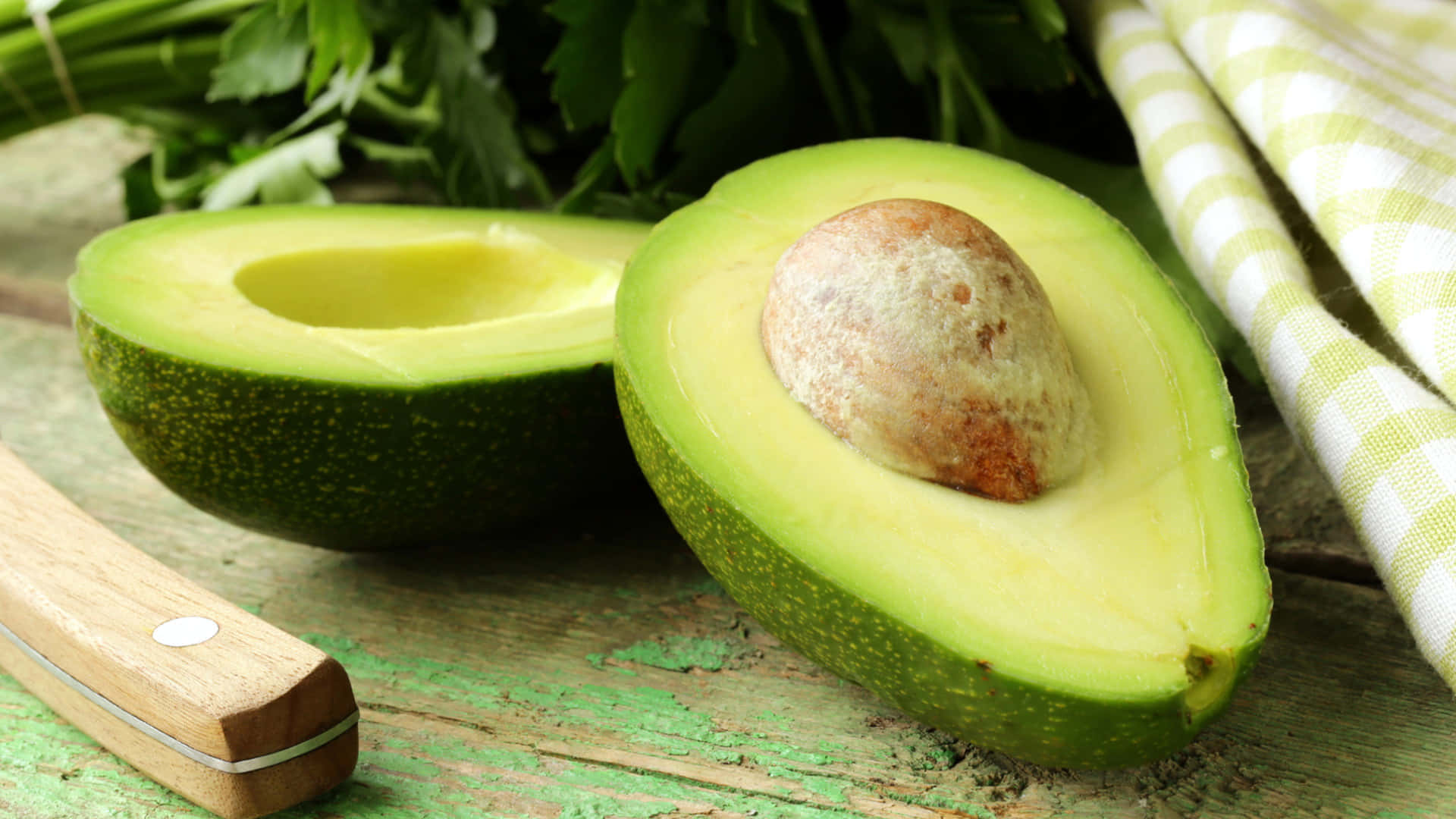 Avocado 桌布 壁紙 — 一個新鮮切開的酪梨，帶有大的棕色果核，放在一個風化的綠色木質表面上，旁邊有歐芹和一塊格子布。
