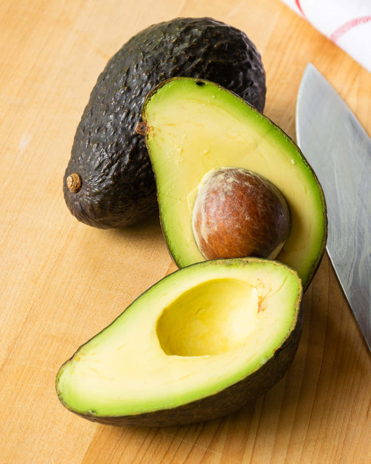 Avocado 桌布 壁紙 — 一個新鮮切開的酪梨露出果核，旁邊放著一個完整的酪梨，在木製砧板上，附近有一把刀。