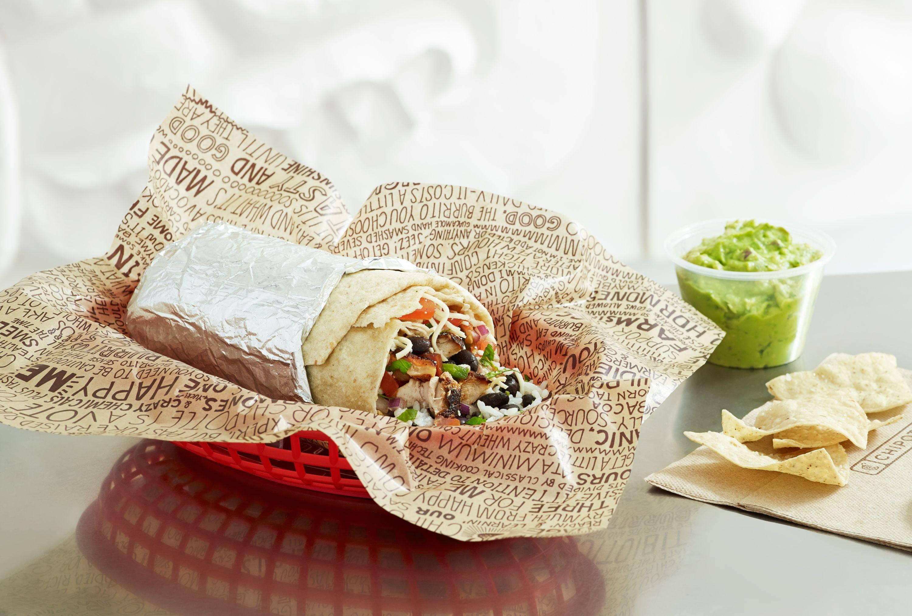 Burrito 桌布 壁紙 — 一個新鮮的墨西哥捲餅，內有黑豆、番茄和酪梨醬，放在紅色籃子裡，旁邊有玉米片和酪梨醬，置於反光桌上。