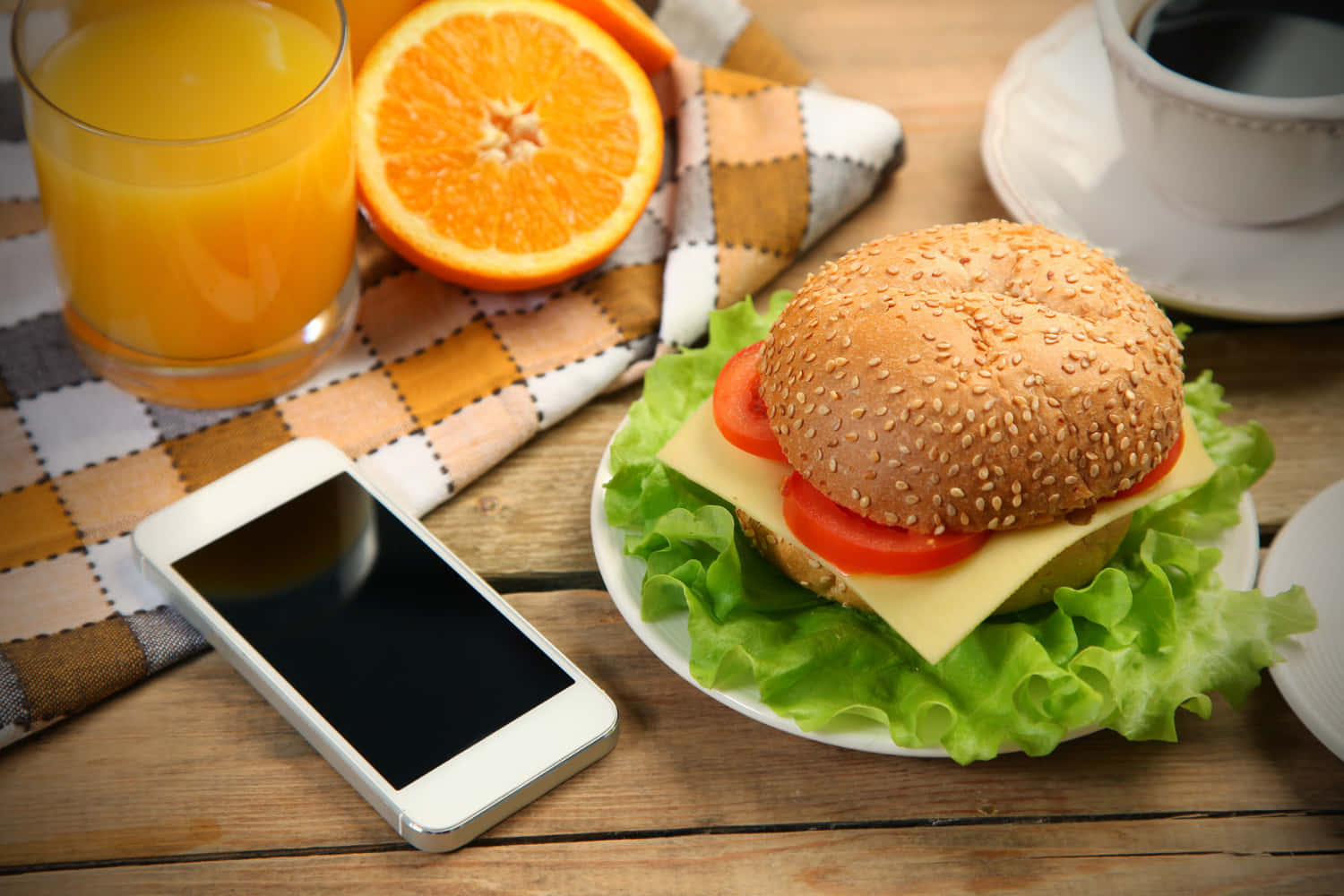 Cheeseburger 桌布 壁紙 — 一個新鮮的芝麻漢堡配生菜、番茄和起司，放在盤子上，旁邊有柳橙汁、切片柳橙和智慧型手機，置於木桌上。