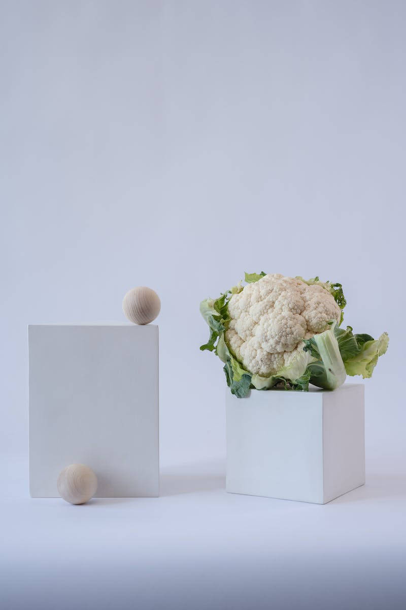 Vegetables 桌布 壁紙 — 一個新鮮的花椰菜，帶有綠葉，放在一個白色立方體中，旁邊是簡約的木球和木塊，背景是柔和的灰色。