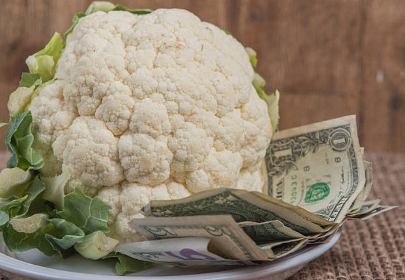 Vegetables 桌布 壁紙 — 一個新鮮的花椰菜連同綠葉放在盤子上，旁邊有散落在編織蓆上的美元紙鈔。