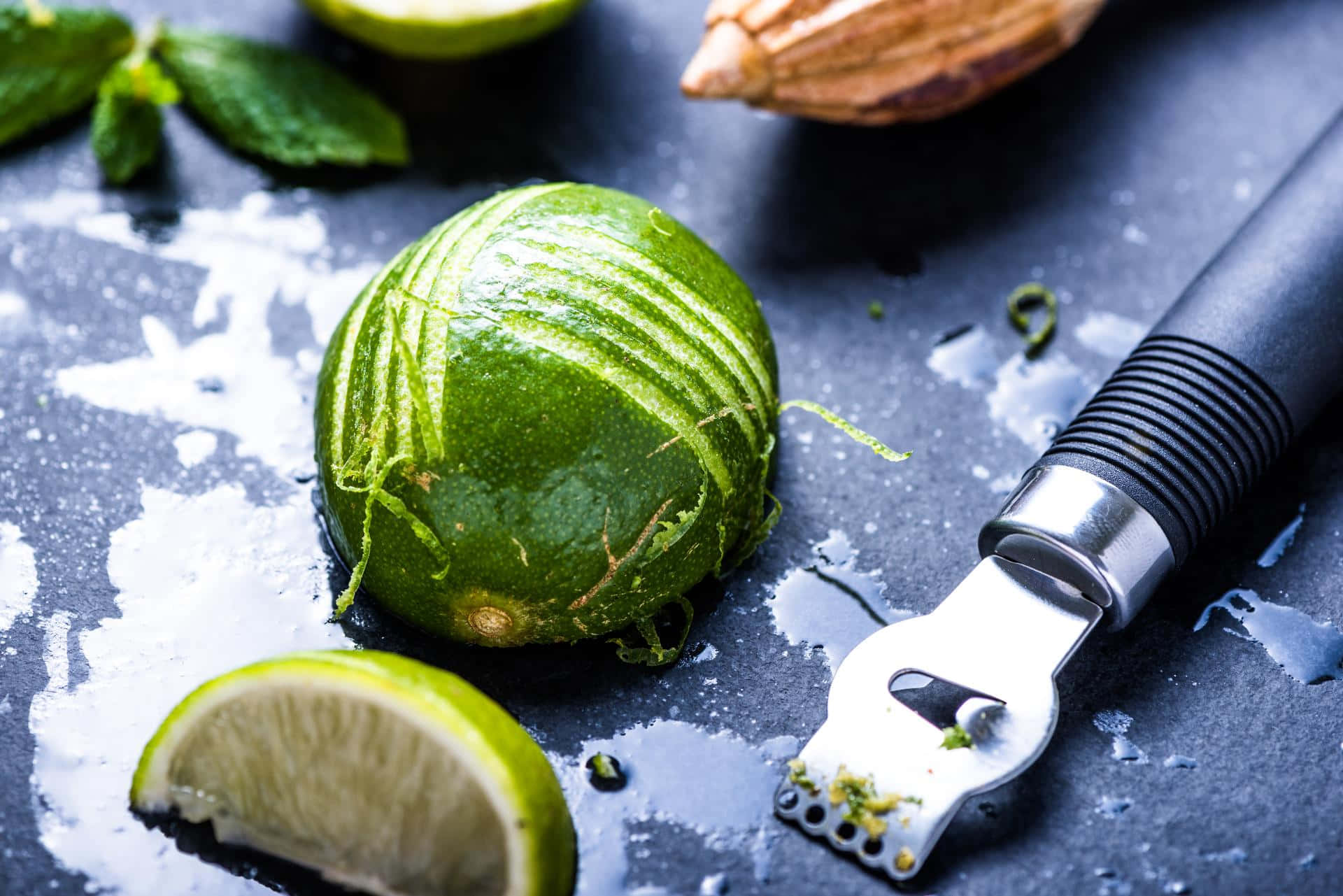 Lime 桌布 壁紙 — 一個新鮮磨皮的萊姆放在一個黑暗潮濕的表面上，旁邊有一個磨皮器和散落的薄荷葉。