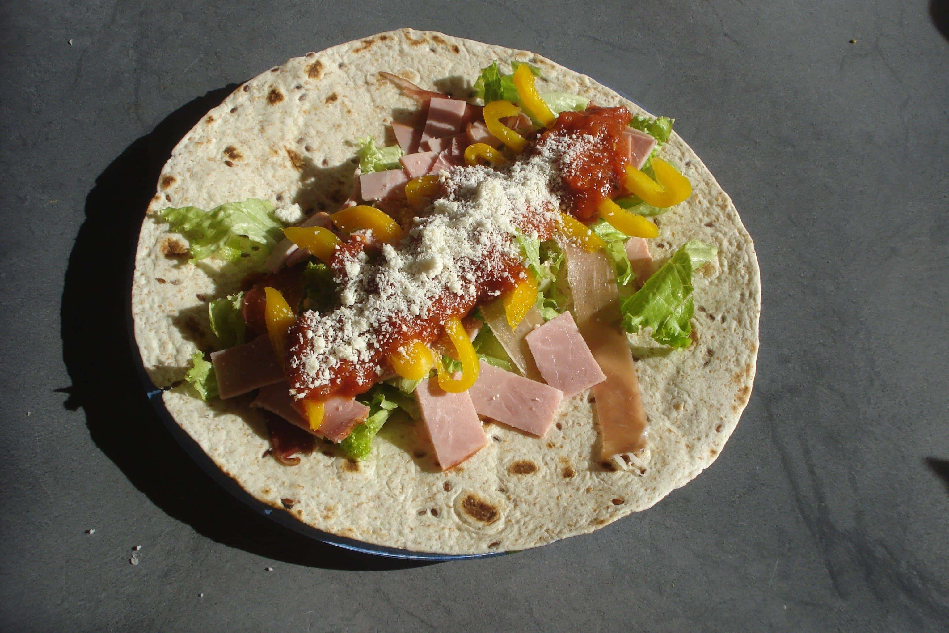 Taco 桌布 壁紙 — 一個新鮮組裝的玉米餅，裡面有火腿、黃椒、生菜和起司，放在深色盤子上，在明亮的陽光下。