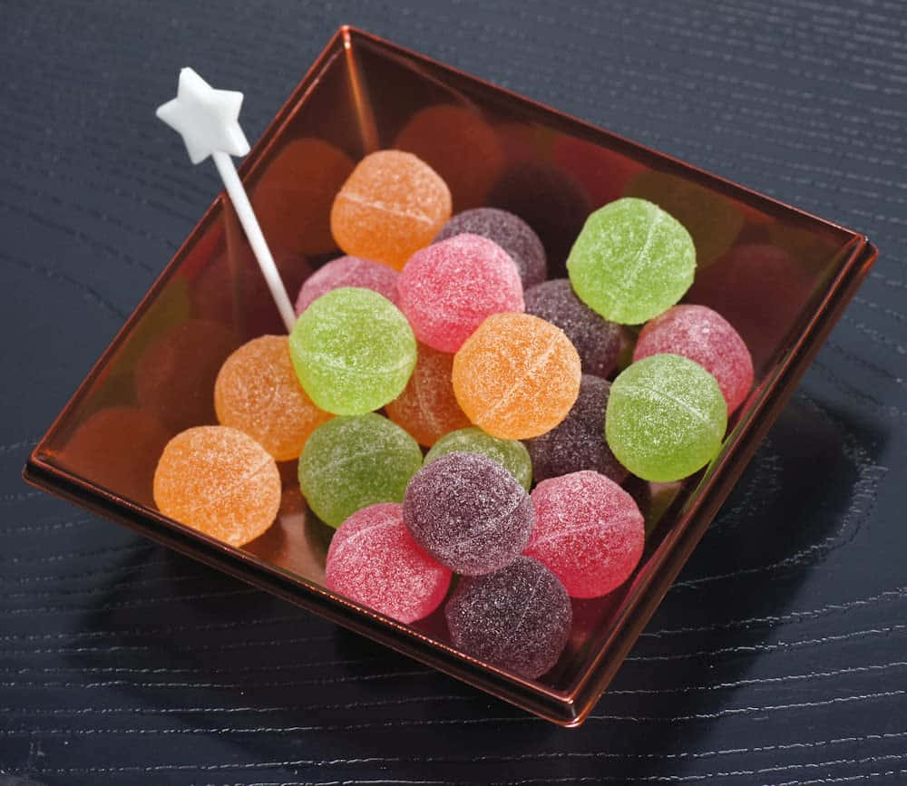 糖果 桌布 壁紙 — 一個方形琥珀色碗，裝滿色彩繽紛的糖衣軟糖和一根白色星星棒，放在深色木質表面上。
