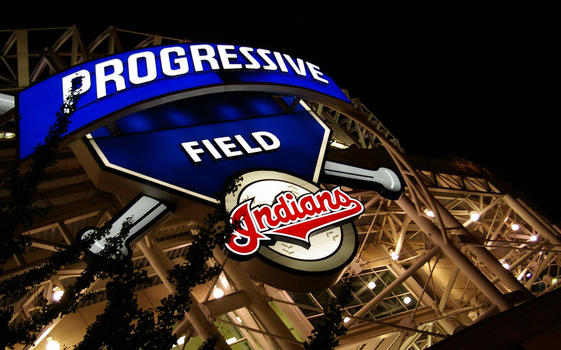 Cleveland Indians 桌布 壁紙 — 一個明亮照明的 Progressive Field 標誌，帶有 Indians 標誌，在黑暗的夜空下發光，由球場建築和樹枝框住。
