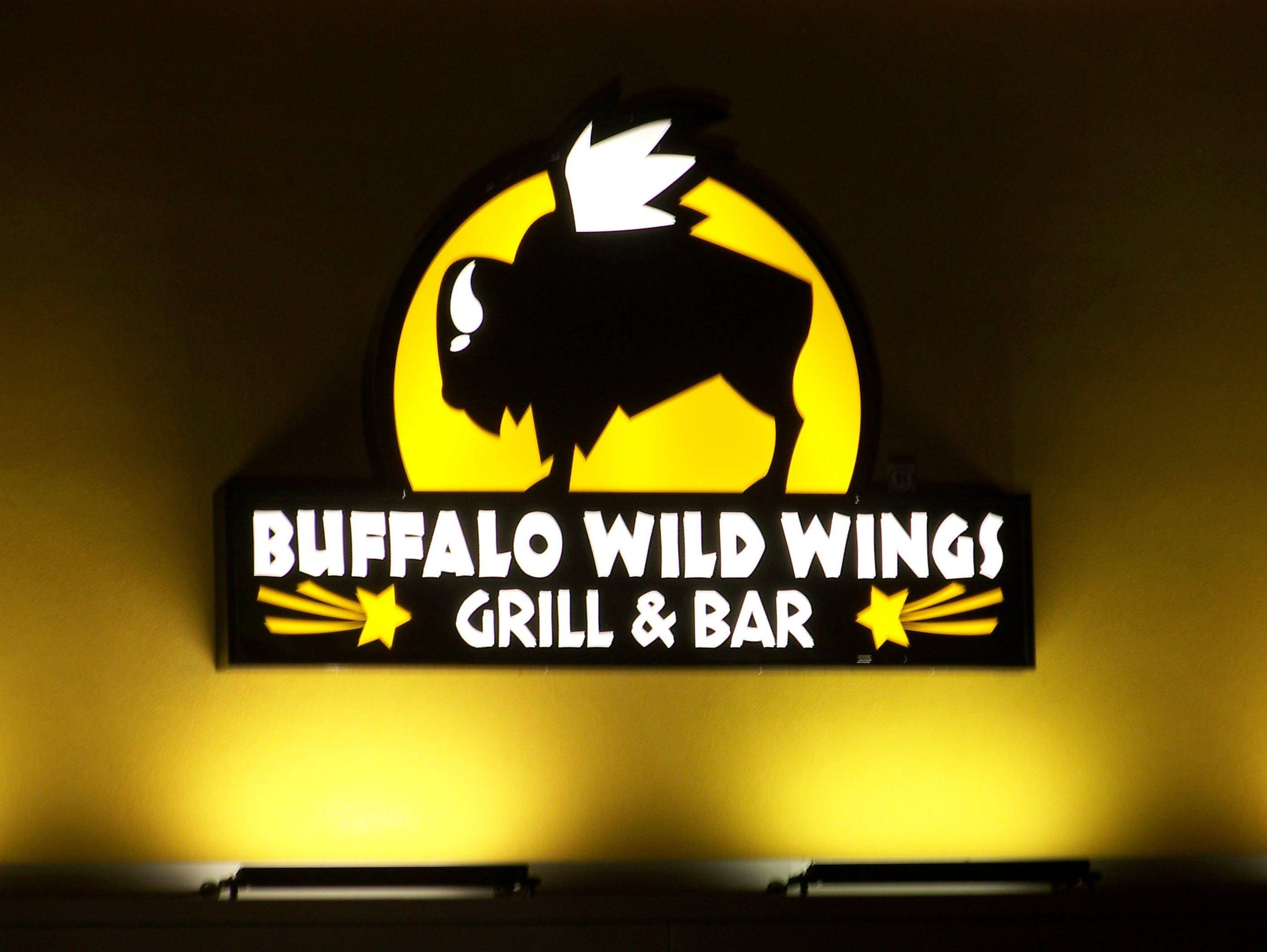 Wild 桌布 壁紙 — 一個明亮照明的Buffalo Wild Wings標誌，黑色野牛剪影映襯在黃色太陽背景上，在牆上投射出溫暖的光芒。