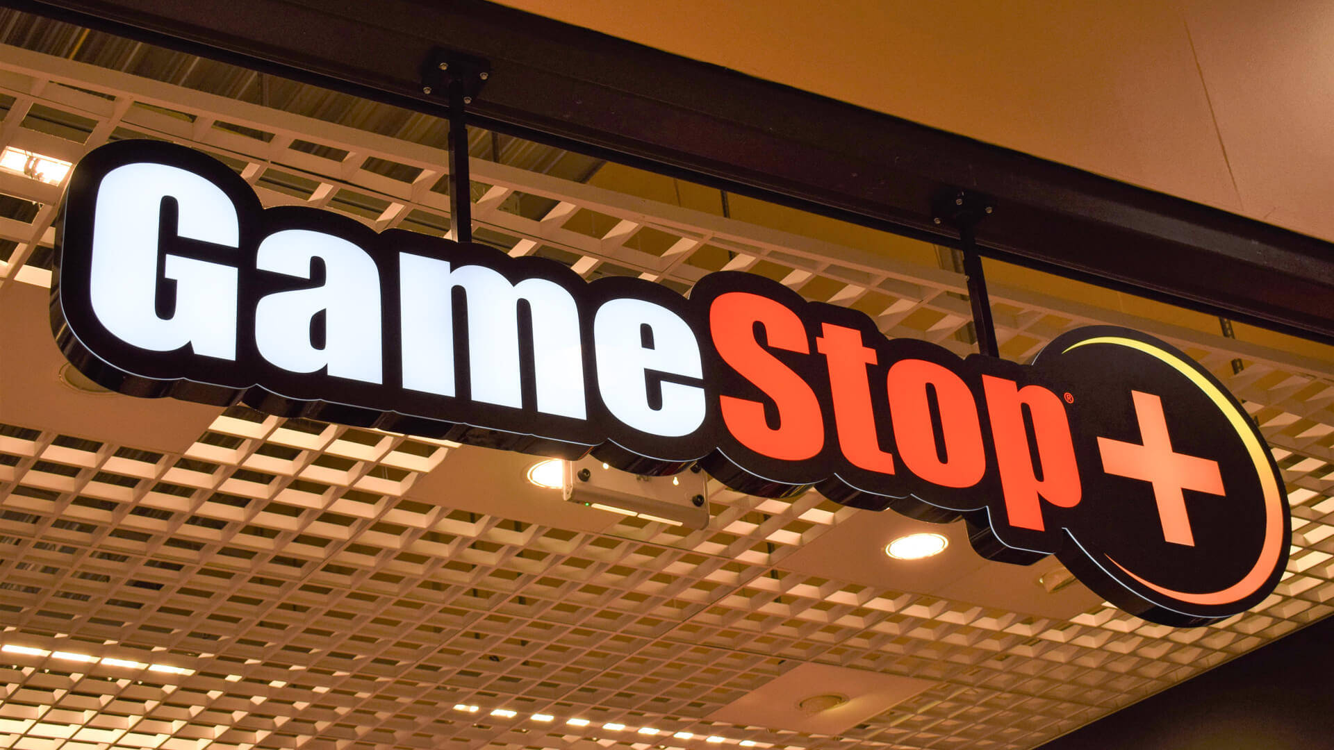 GameStop 桌布 壁紙 — 一個明亮照明的GameStop Plus標誌懸掛在溫暖照明的室內空間的網格天花板上。