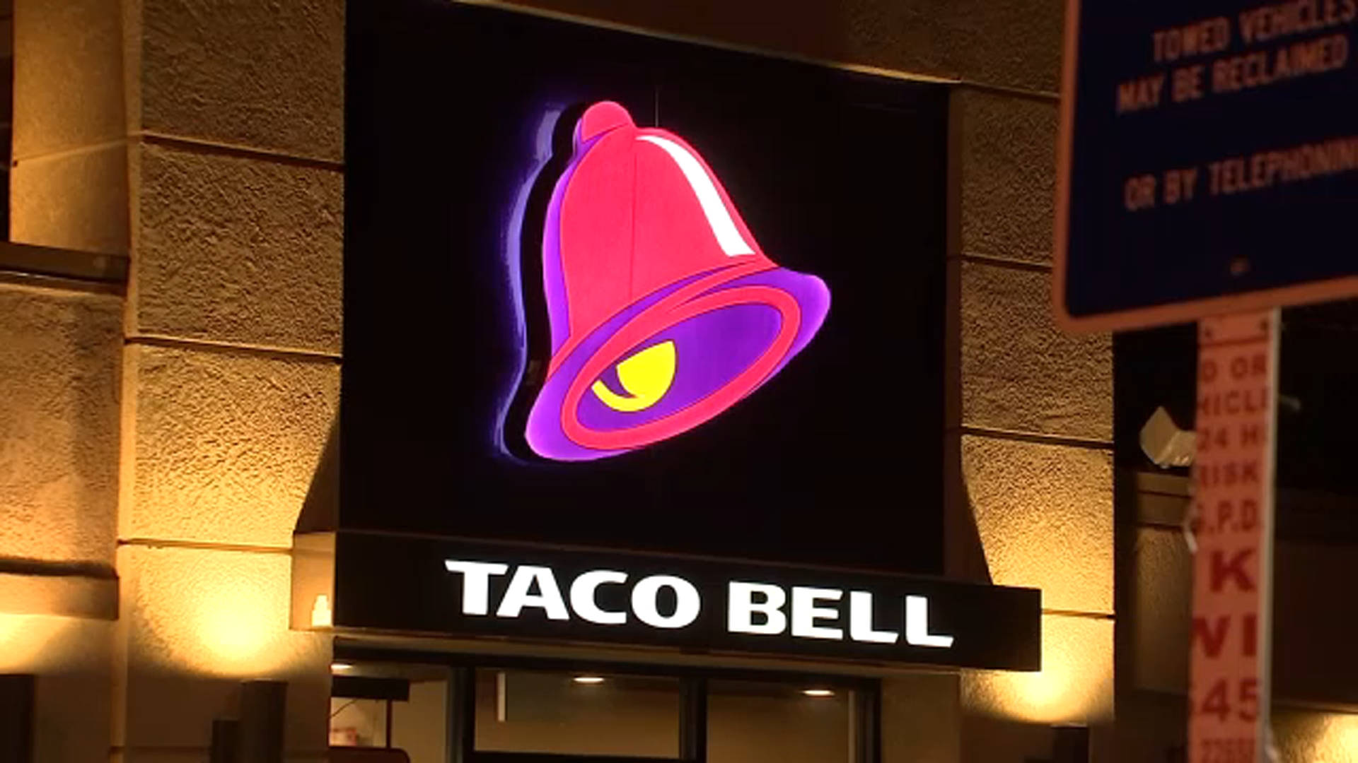 塔可鐘 桌布 壁紙 — 一個明亮照明的Taco Bell招牌，帶有發光的粉紅色和紫色鈴鐺標誌，在石頭建築外牆上方的黑暗夜空中閃耀。