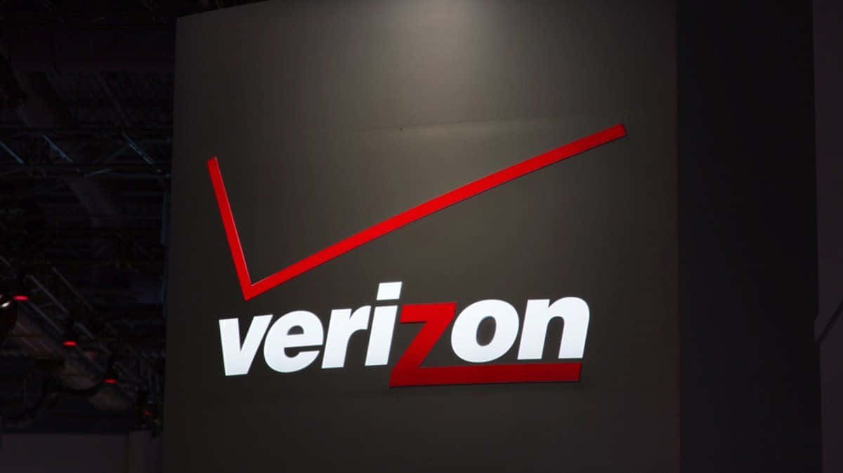 威訊 桌布 壁紙 — 一個明亮照明的Verizon標誌，帶有紅色和白色字樣，位於昏暗室內環境中的深灰色牆壁上。