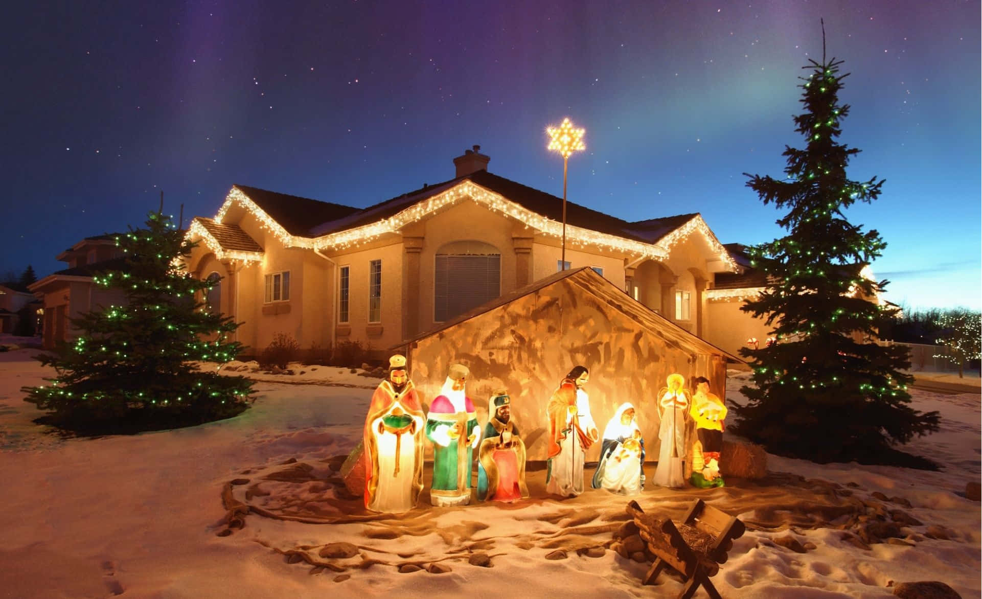 Nativity 桌布 壁紙 — 一個明亮照明的戶外耶穌誕生場景，帶有發光的人物，矗立在雪覆蓋的房子前，在星空下。