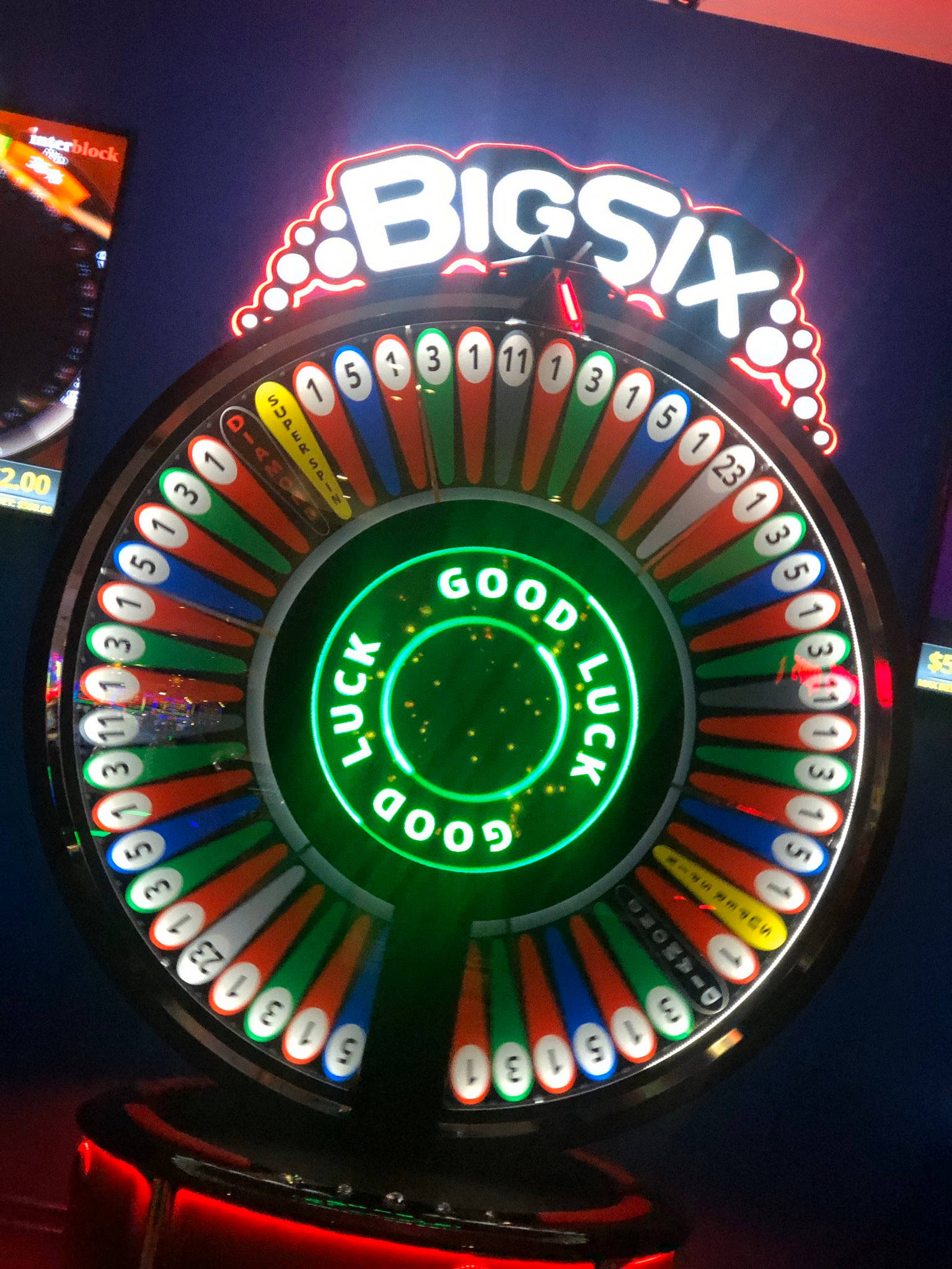 霓虹燈 桌布 壁紙 — 一個明亮照明的賭場輪盤，標有「BIG SIX」，中心有發光的綠色「GOOD LUCK」字樣和彩色編號的扇區。