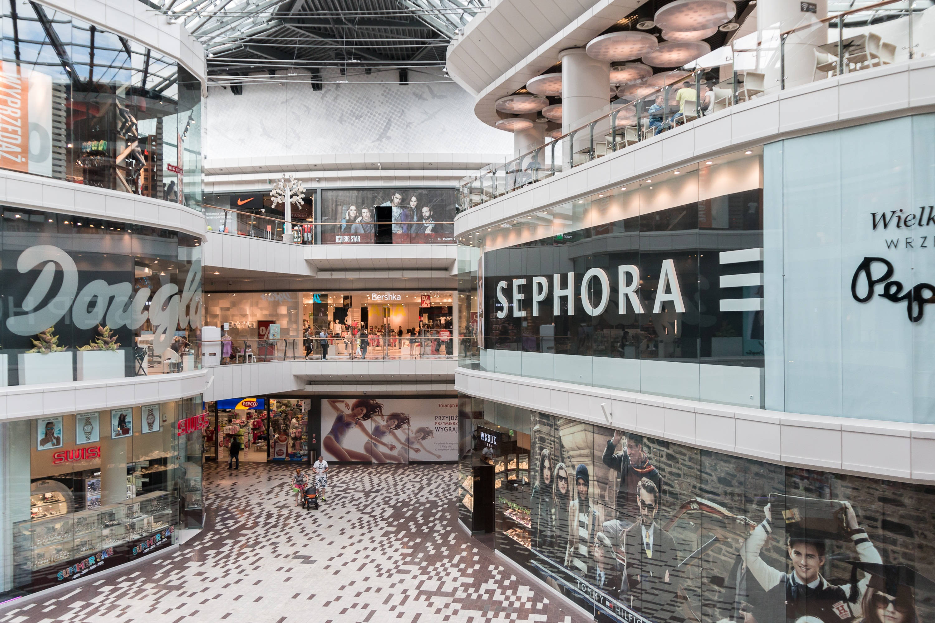 Mall 桌布 壁紙 — 一個明亮的多層購物中心，可見Sephora和Dough等商店，設有玻璃欄杆和圖案地板，高天花板下。