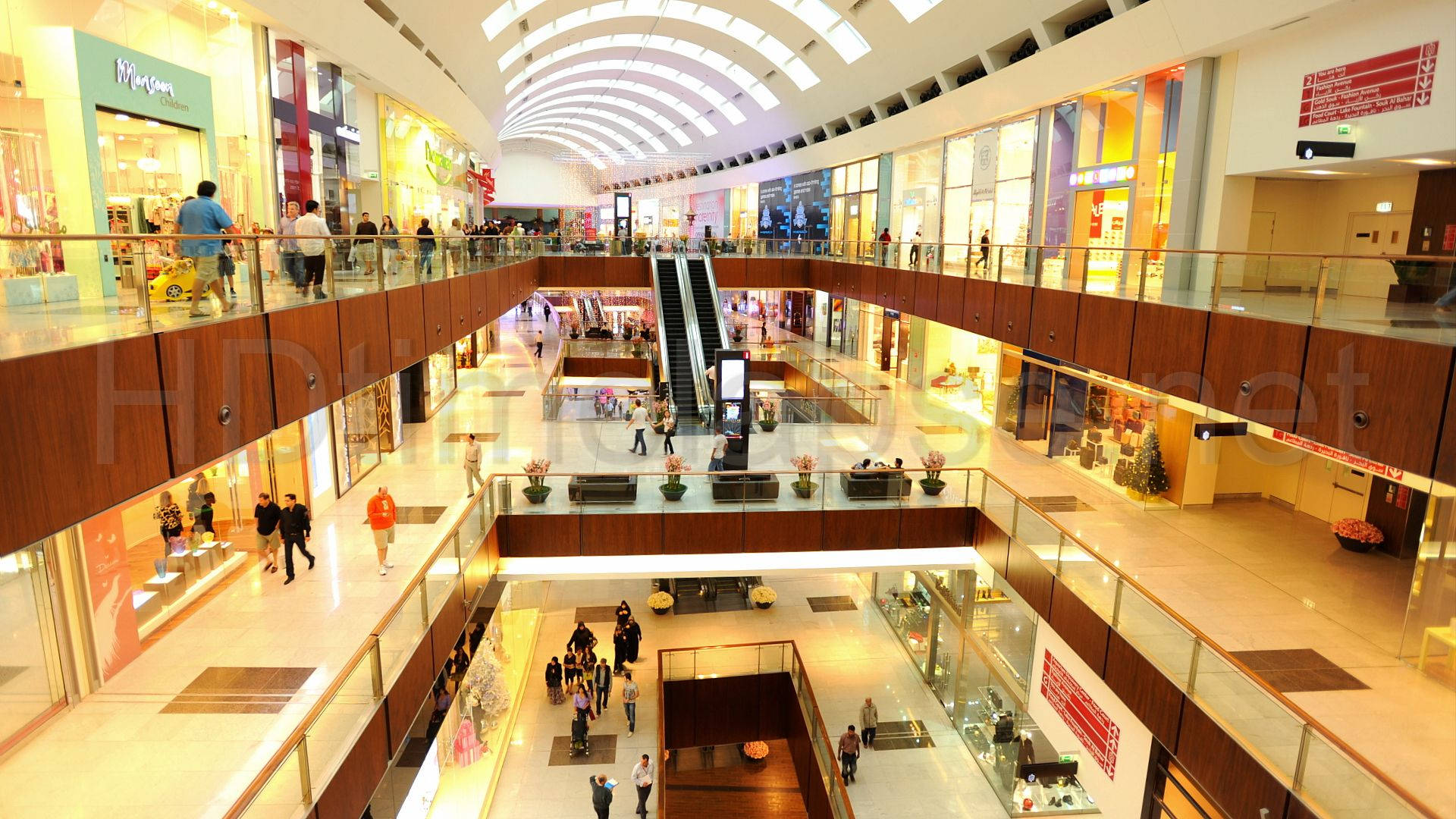 Shopping 桌布 壁紙 — 一個明亮的多層購物中心，設有玻璃欄杆、木質裝飾，購物者在弧形天窗下穿梭於商店之間。