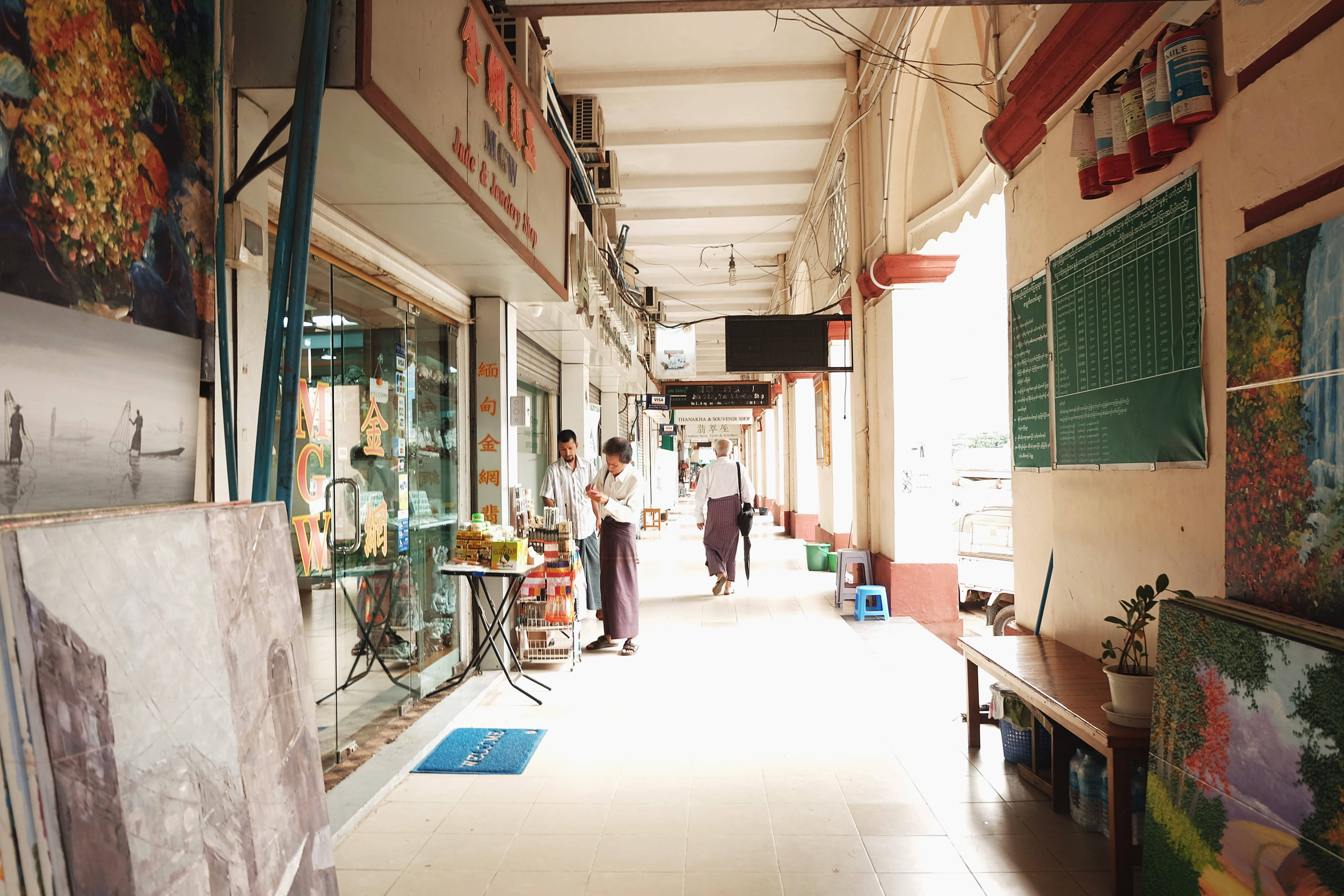 Yangon 桌布 壁紙 — 一個明亮的帶頂棚小巷，在熙熙攘攘的市場中，有攤販和購物者在溫暖的陽光下。