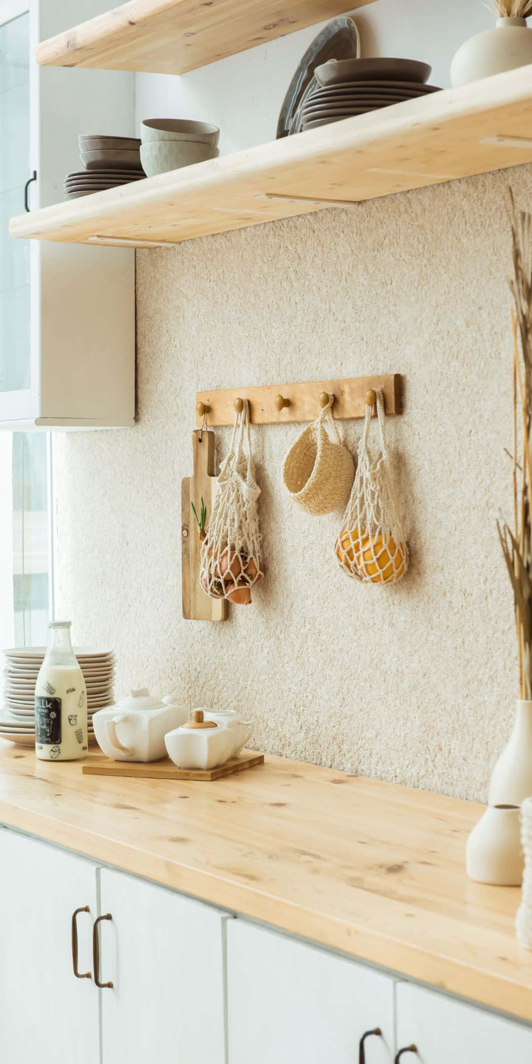 Pixel 3 Kitchen 桌布 壁紙 — 一個明亮的極簡主義廚房，淺色木架上放著疊放的盤子，掛著編織袋裝著農產品，背景是帶紋理的米色牆壁。
