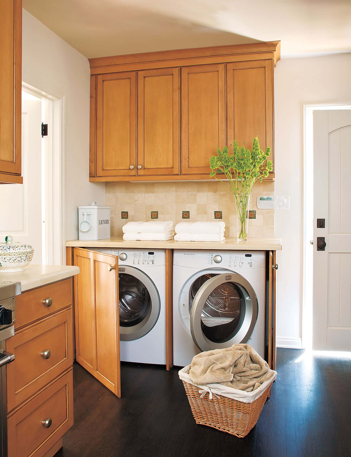 laundry 桌布 壁紙 — 一個明亮的洗衣房，配有木製櫥櫃、白色電器和一個裝有摺疊毛巾的柳條籃。