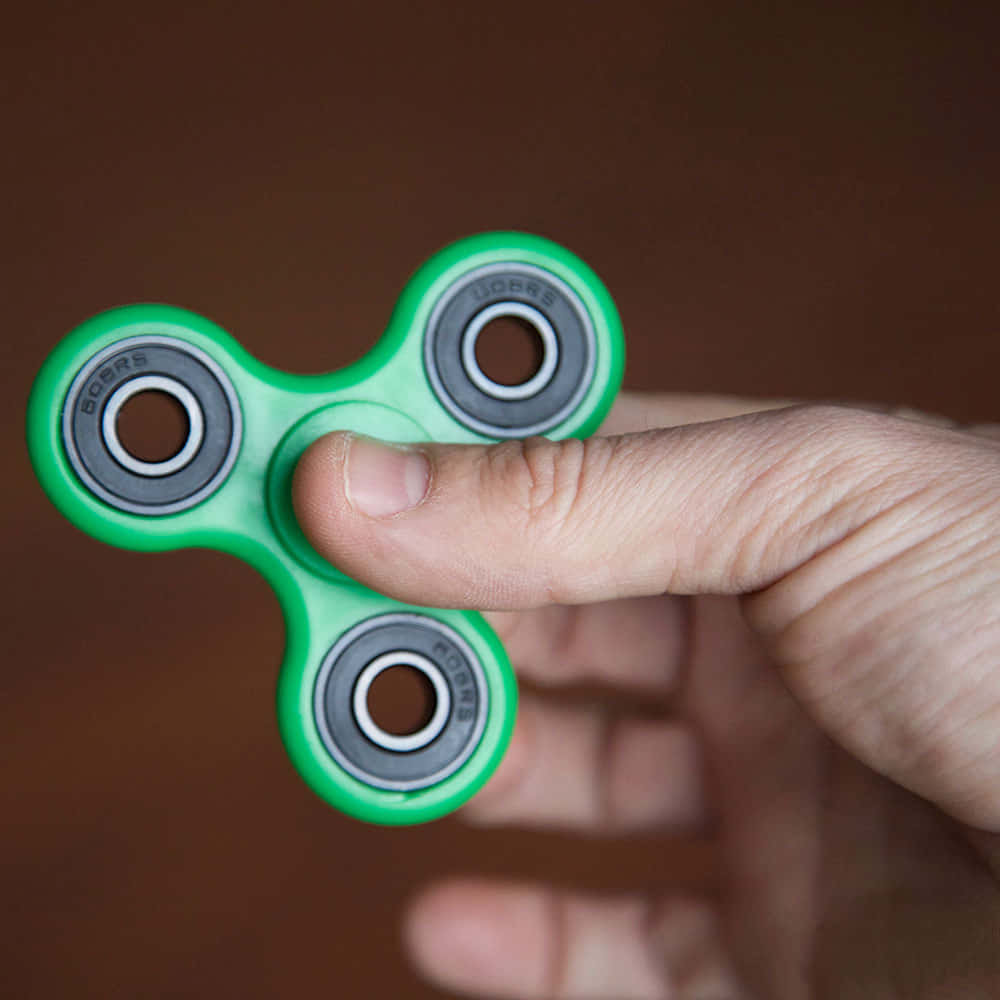 Fidget 桌布 壁紙 — 一個明亮的綠色指尖陀螺被手指拿著的近距離特寫，背景是模糊的棕色。