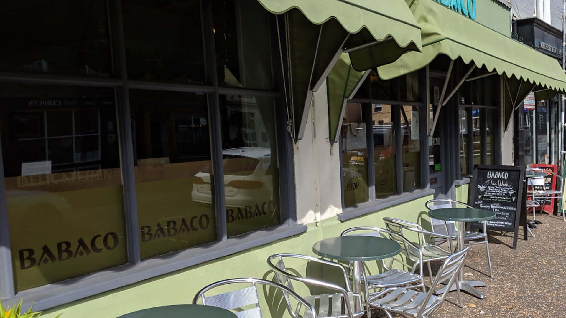 Restaurant 桌布 壁紙 — 一個明亮的綠色遮陽篷為名為 Babaco 的咖啡館的戶外桌椅提供遮蔭，大窗戶反射著街道。