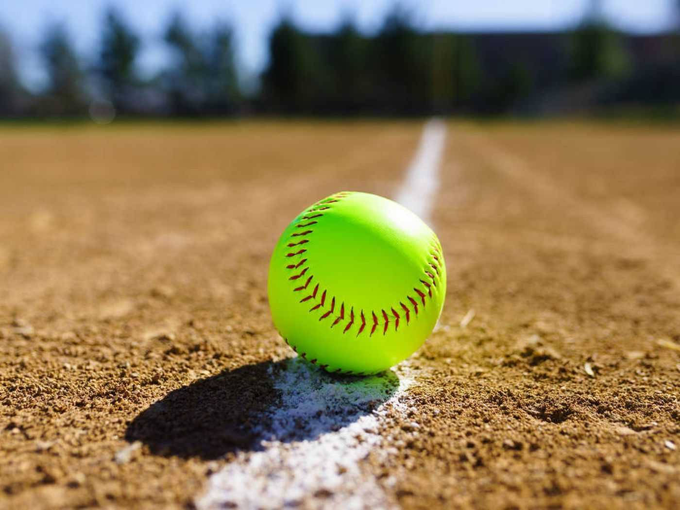 Cute Softball 桌布 壁紙 — 一個明亮的螢光黃色壘球放在陽光照耀的泥地球場上，後面有一條白色粉筆線。
