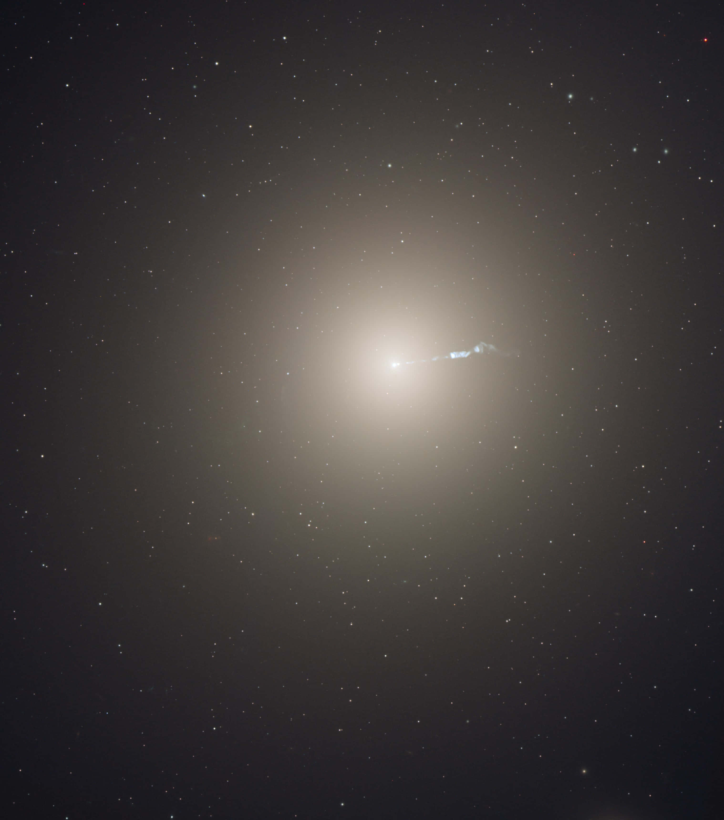 Elliptical Galaxy 桌布 壁紙 — 一個明亮的螺旋星系在星場中柔和地發光，其核心延伸出一條微弱的藍色噴流。