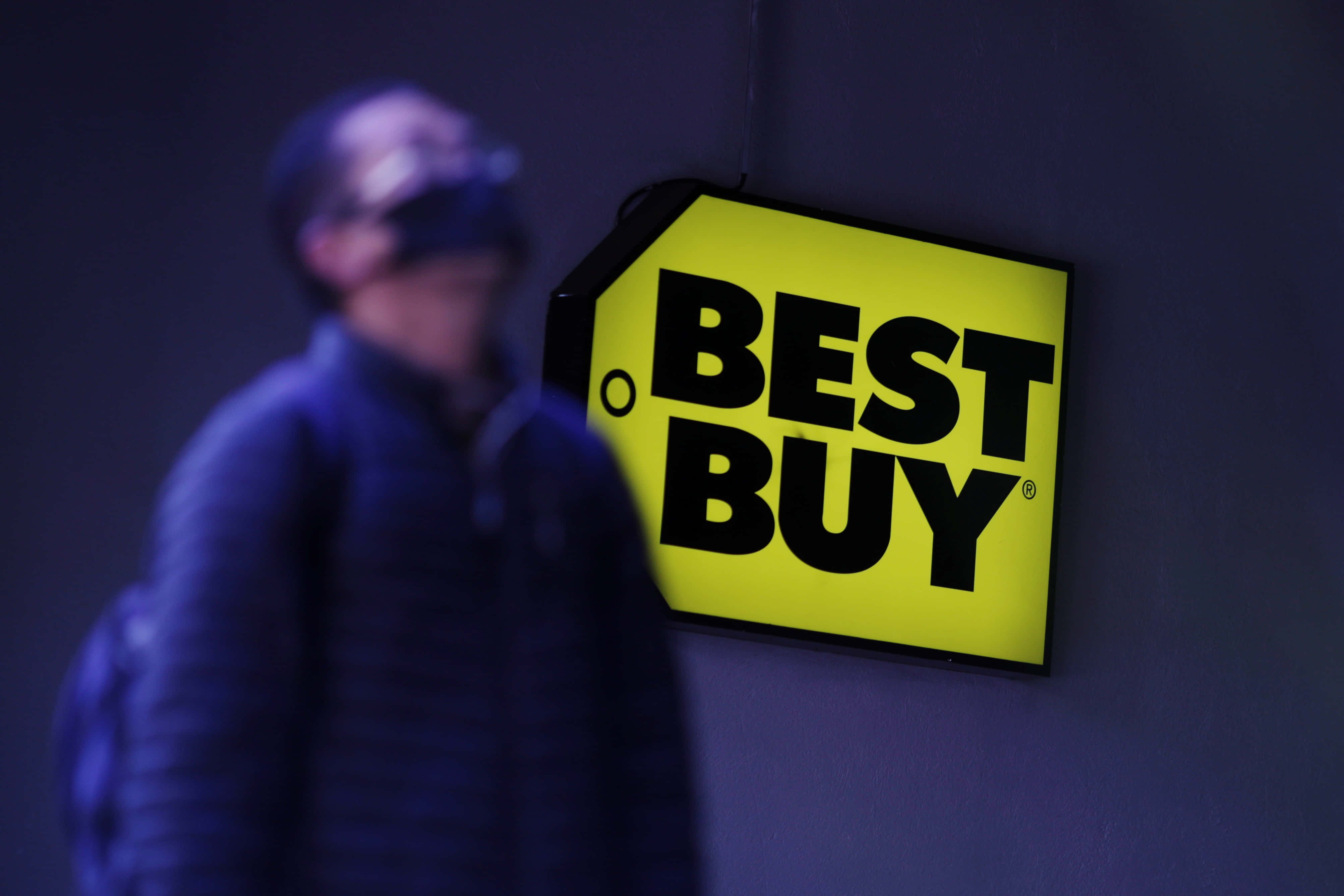 Best Buy 桌布 壁紙 — 一個明亮的黃色Best Buy標誌在黑暗的牆壁上發光，一個模糊的人影在陰暗的藍色調中走過。