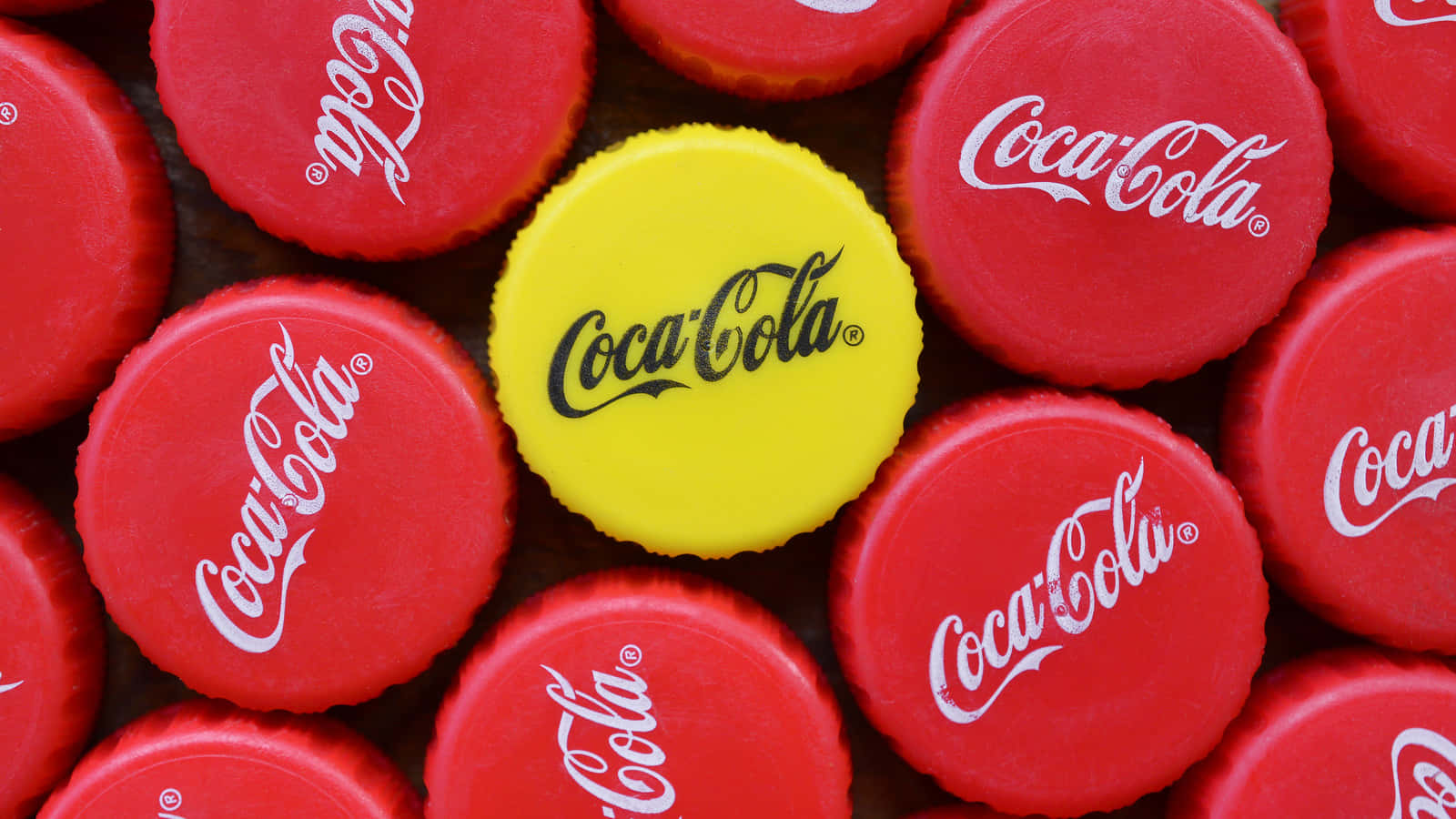 coca cola 桌布 壁紙 — 一個明亮的黃色可口可樂瓶蓋，被紅色瓶蓋包圍在深色表面上。