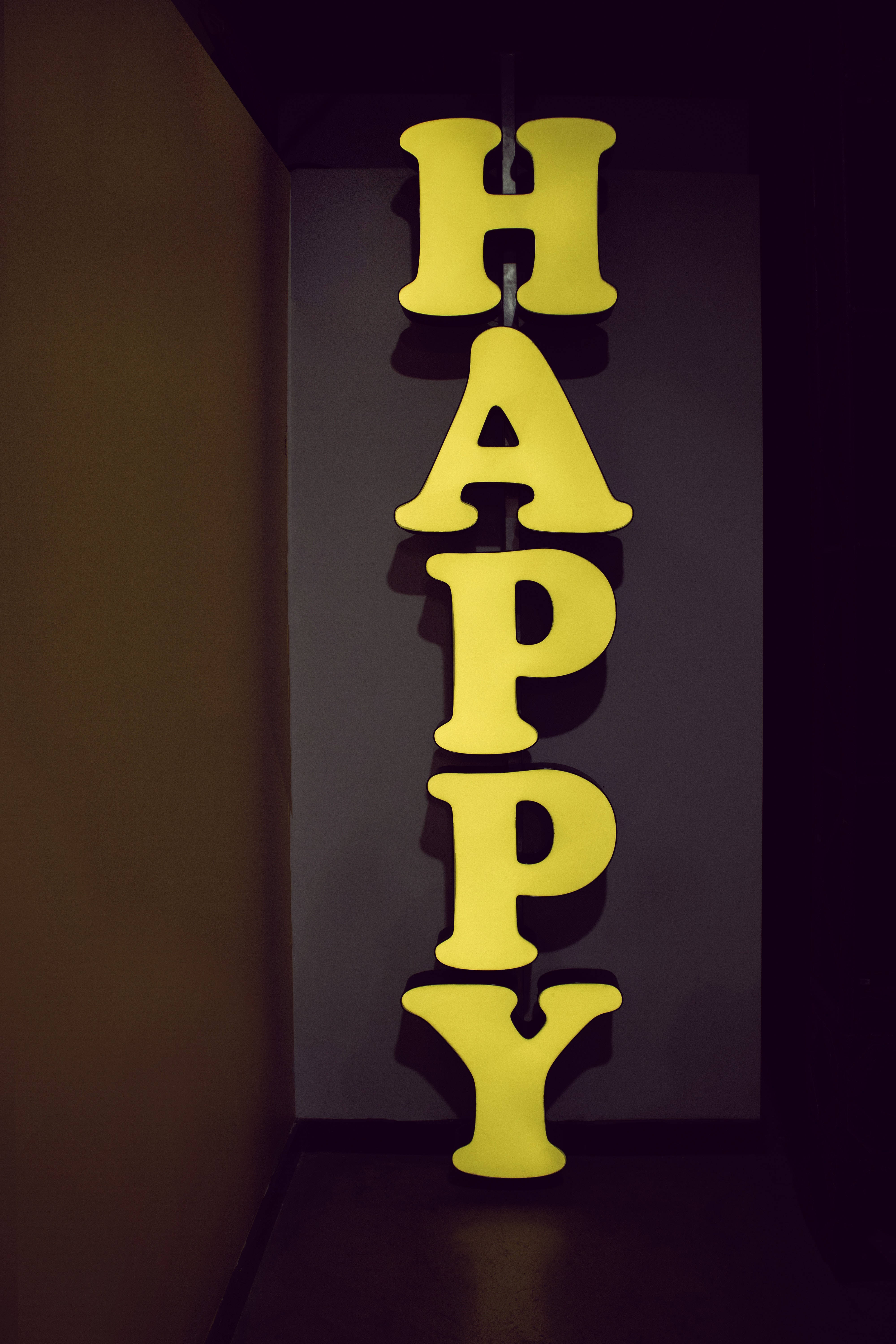 桌布 壁紙 — 一個明亮的黃色垂直霓虹燈牌，拼寫著HAPPY，在昏暗的角落裡對著深色牆壁溫暖地發光。