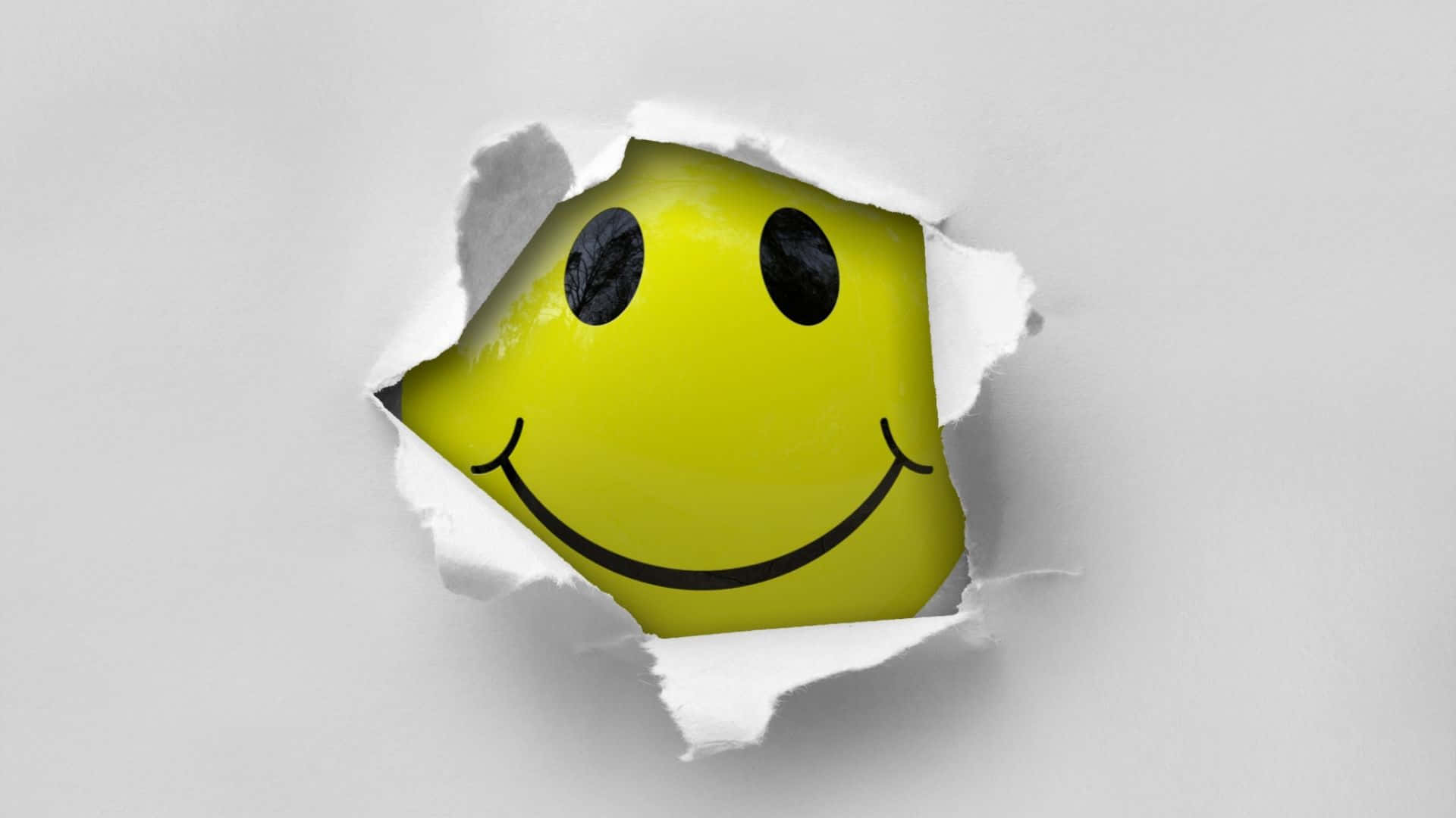 Smiley 桌布 壁紙 — 一個明亮的黃色笑臉從一個邊緣參差不齊的撕破白色紙洞中探出頭來。