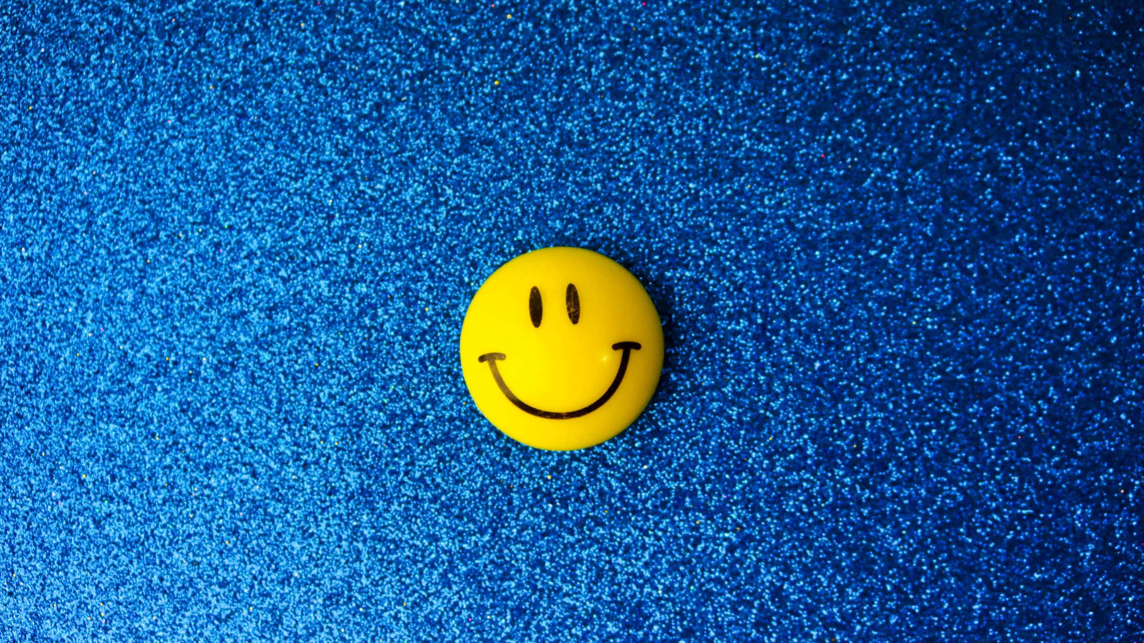 Smile 桌布 壁紙 — 一個明亮的黃色笑臉按鈕放置在閃閃發光的藍色亮粉背景上。
