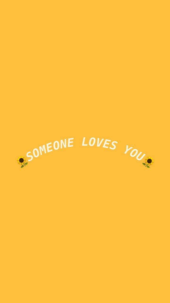 桌布 壁紙 — 一個明亮的黃色背景，白色文字「someone loves you」在兩個向日葵表情符號之間彎曲排列。