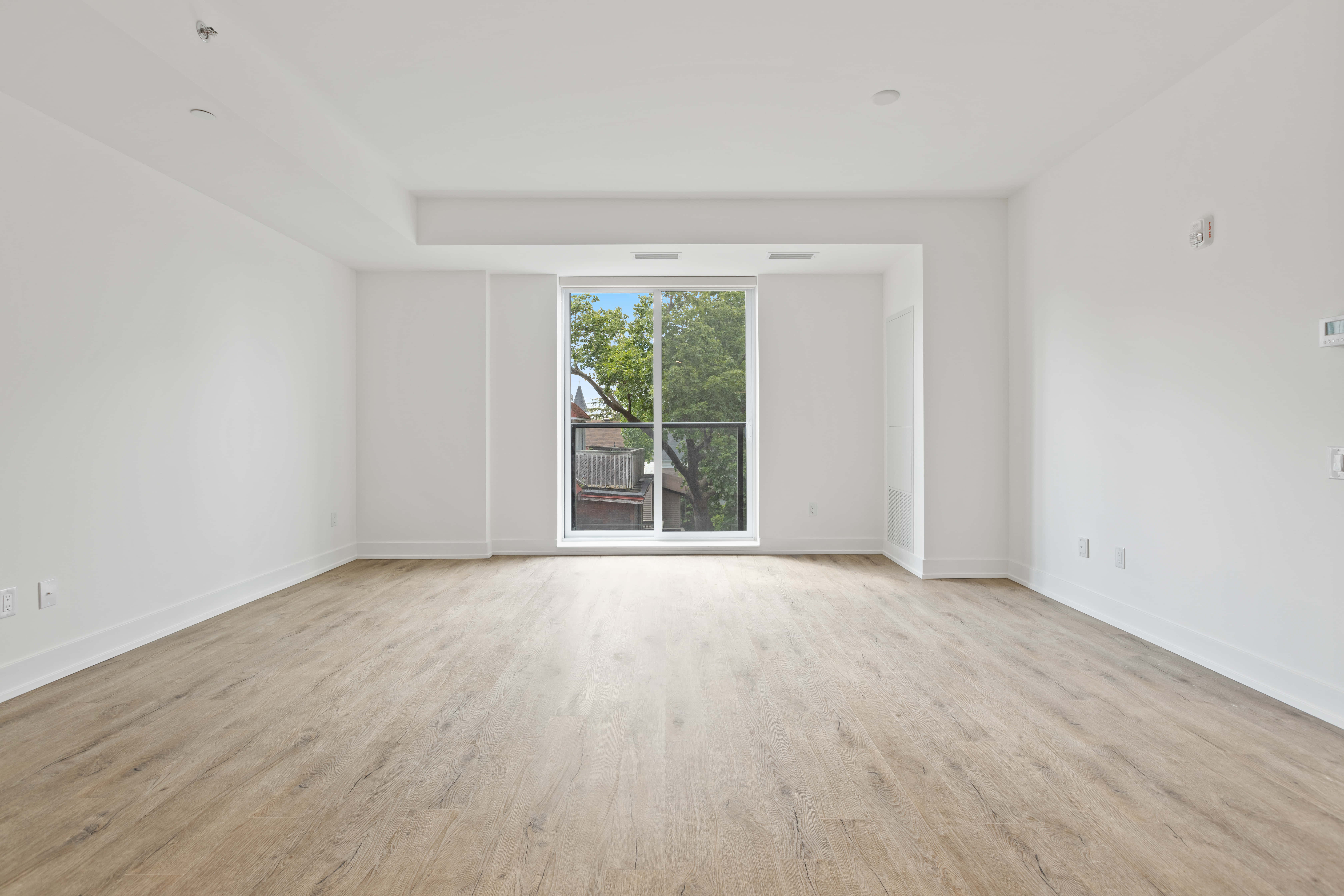 Empty Room 桌布 壁紙 — 一個明亮簡約的空房間，鋪有淺色木地板和白色牆壁，大窗戶展現出外面的綠色樹木。