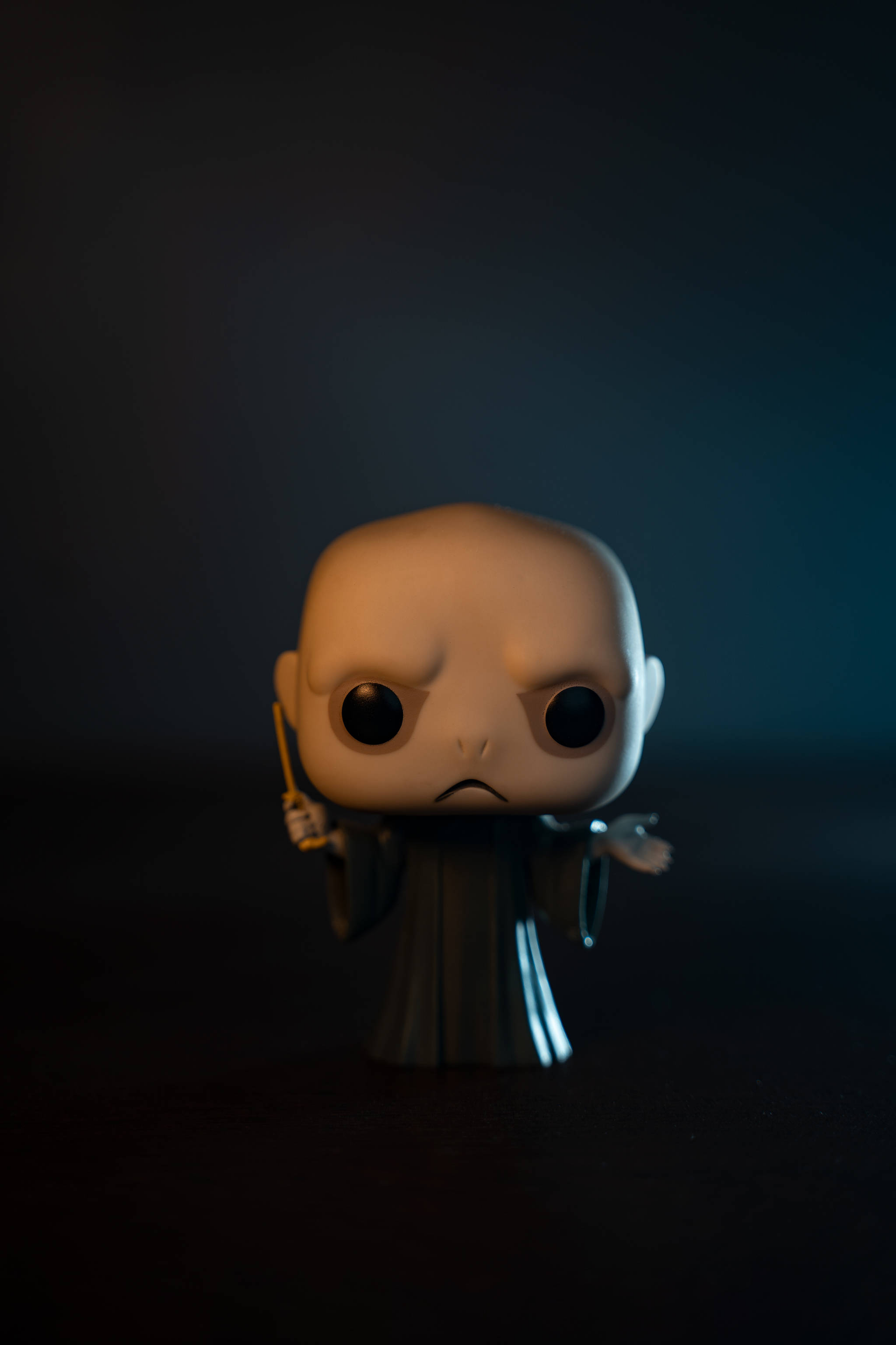 pop 桌布 壁紙 — 一個昏暗燈光下的佛地魔Funko Pop公仔，表情嚴肅，手持魔杖，背景為深色漸變。
