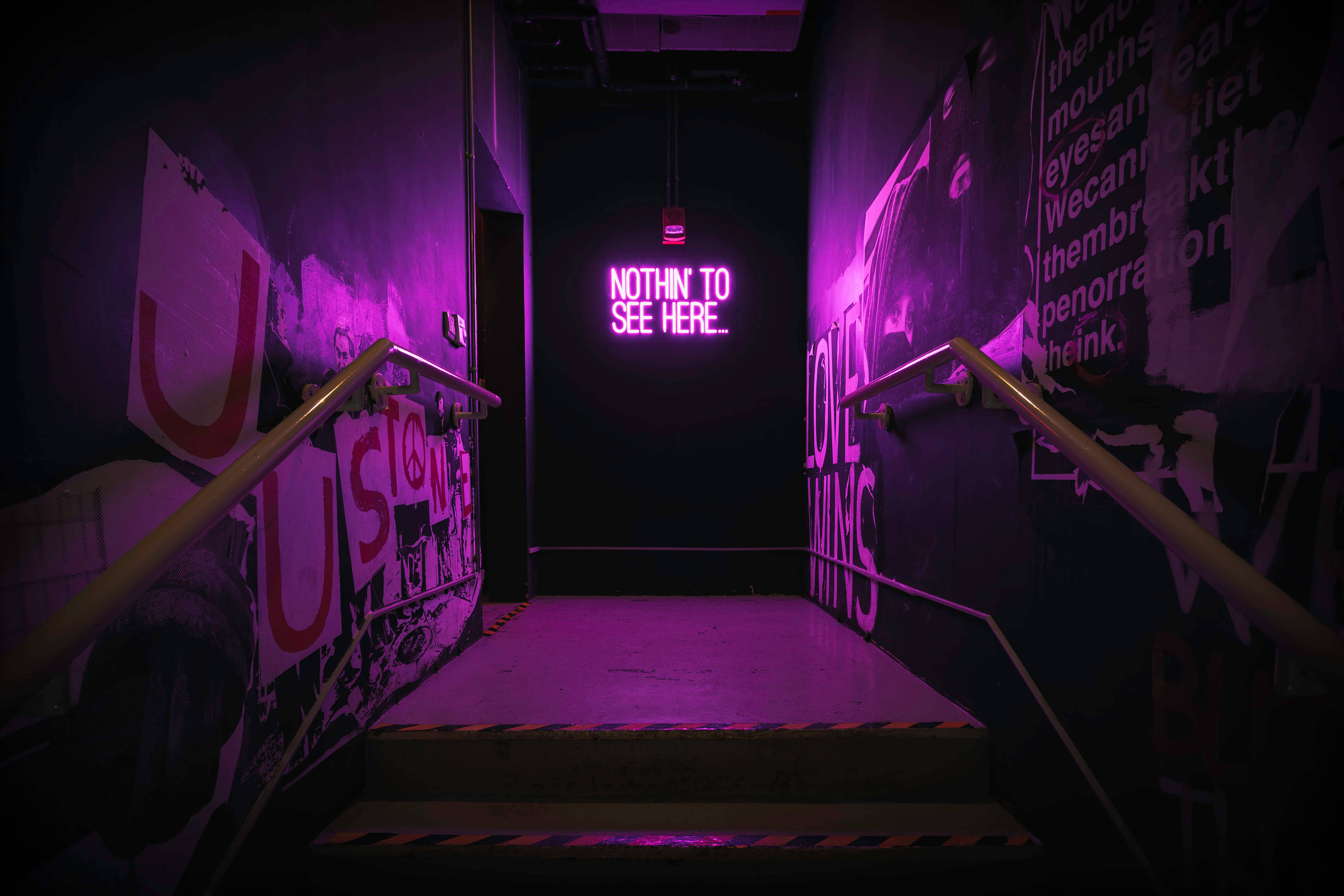 Dark Neon 桌布 壁紙 — 一個昏暗的樓梯間，沐浴在紫色霓虹燈光中，牆壁佈滿塗鴉，還有一個發光的標誌寫著「NOTHIN' TO SEE HERE。」