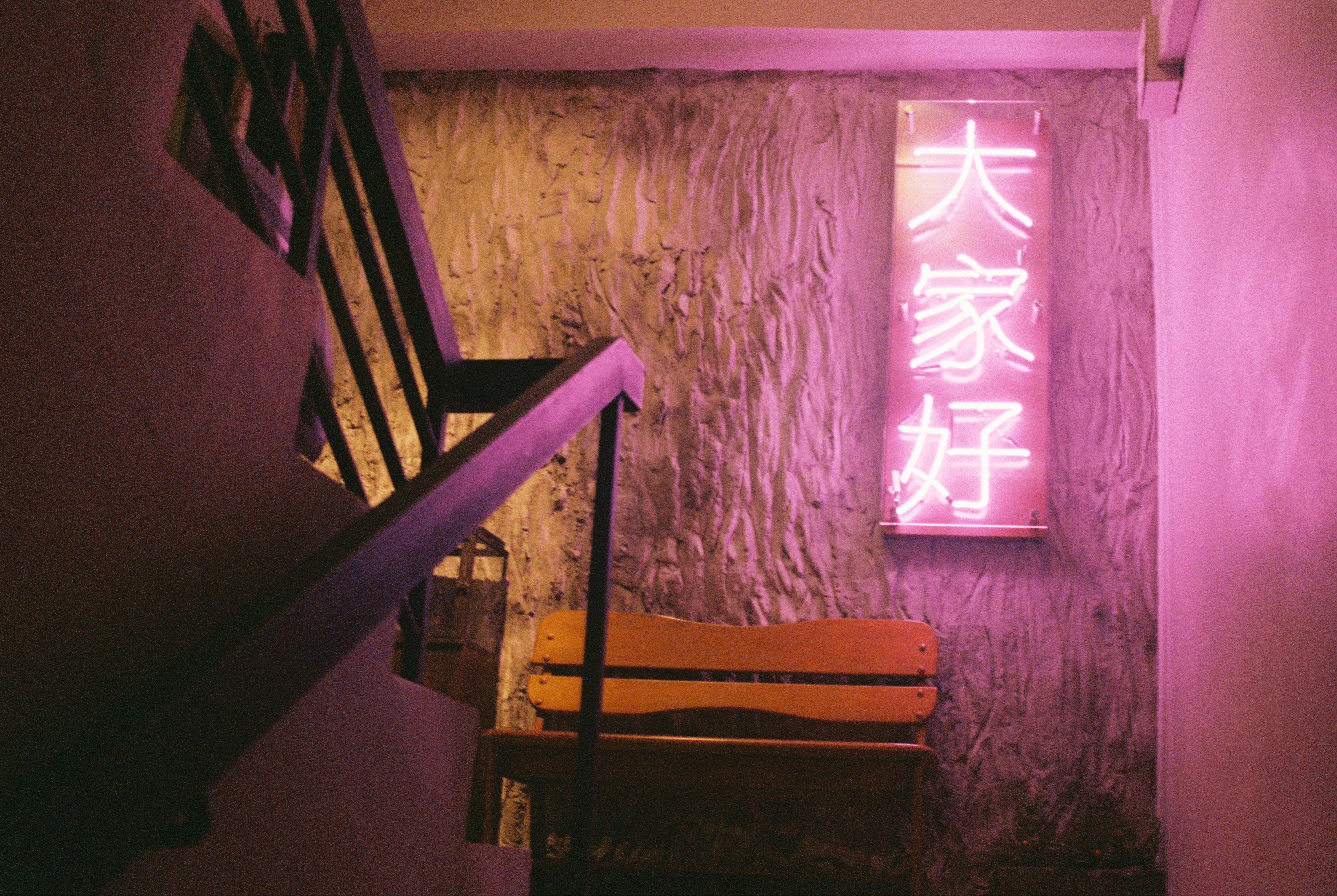 Aesthetic Photography 桌布 壁紙 — 一個昏暗的樓梯間，被一個帶有中文字的垂直標誌的粉紅色霓虹燈光沐浴，在紋理牆壁和木長凳上投射出柔和的光線。