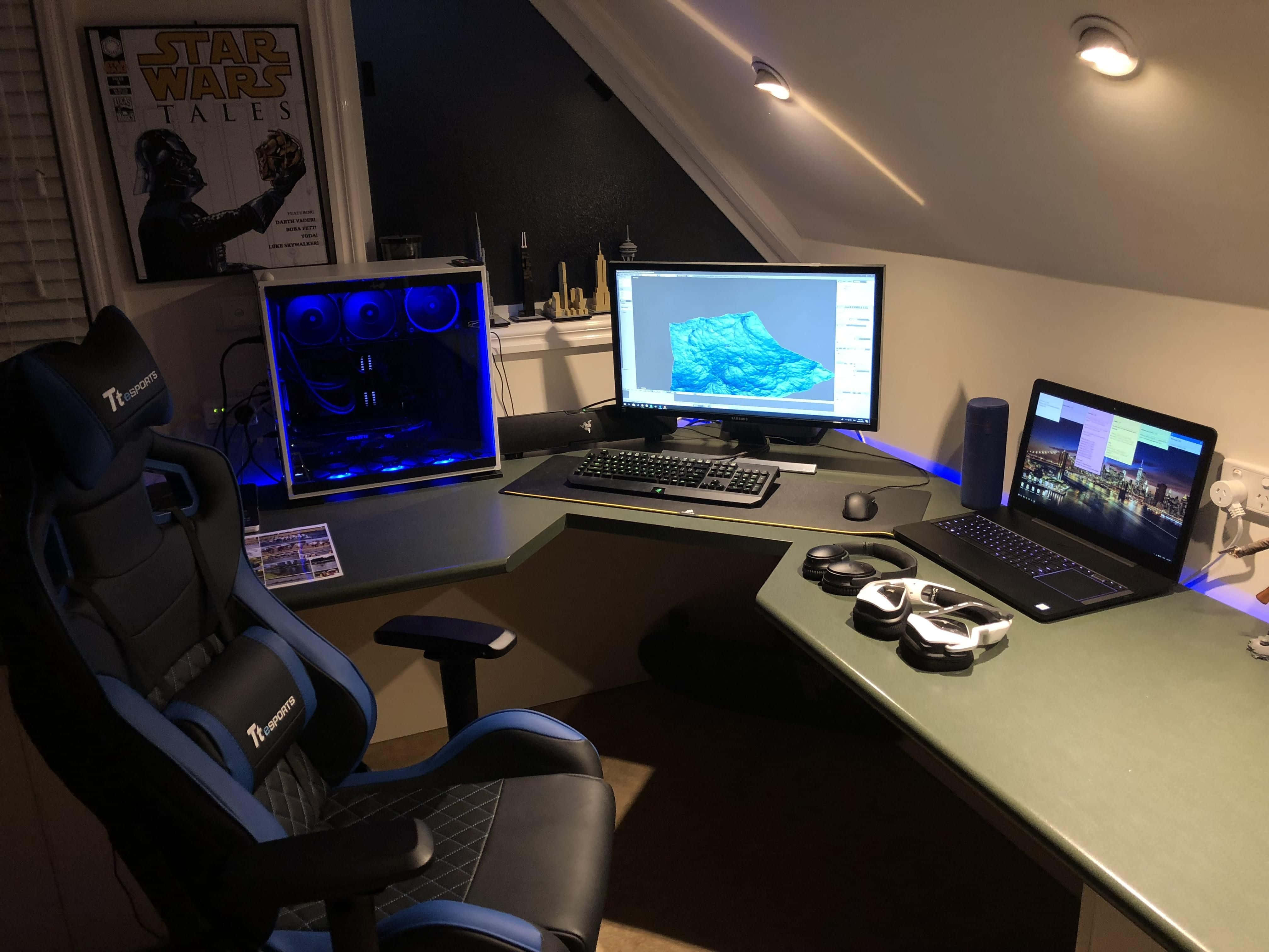 Gaming Pc Setup 桌布 壁紙 — 一個昏暗的遊戲設置，配有藍色發光的電腦主機、多個顯示器和一張賽車椅，在溫暖的天花板燈光下。