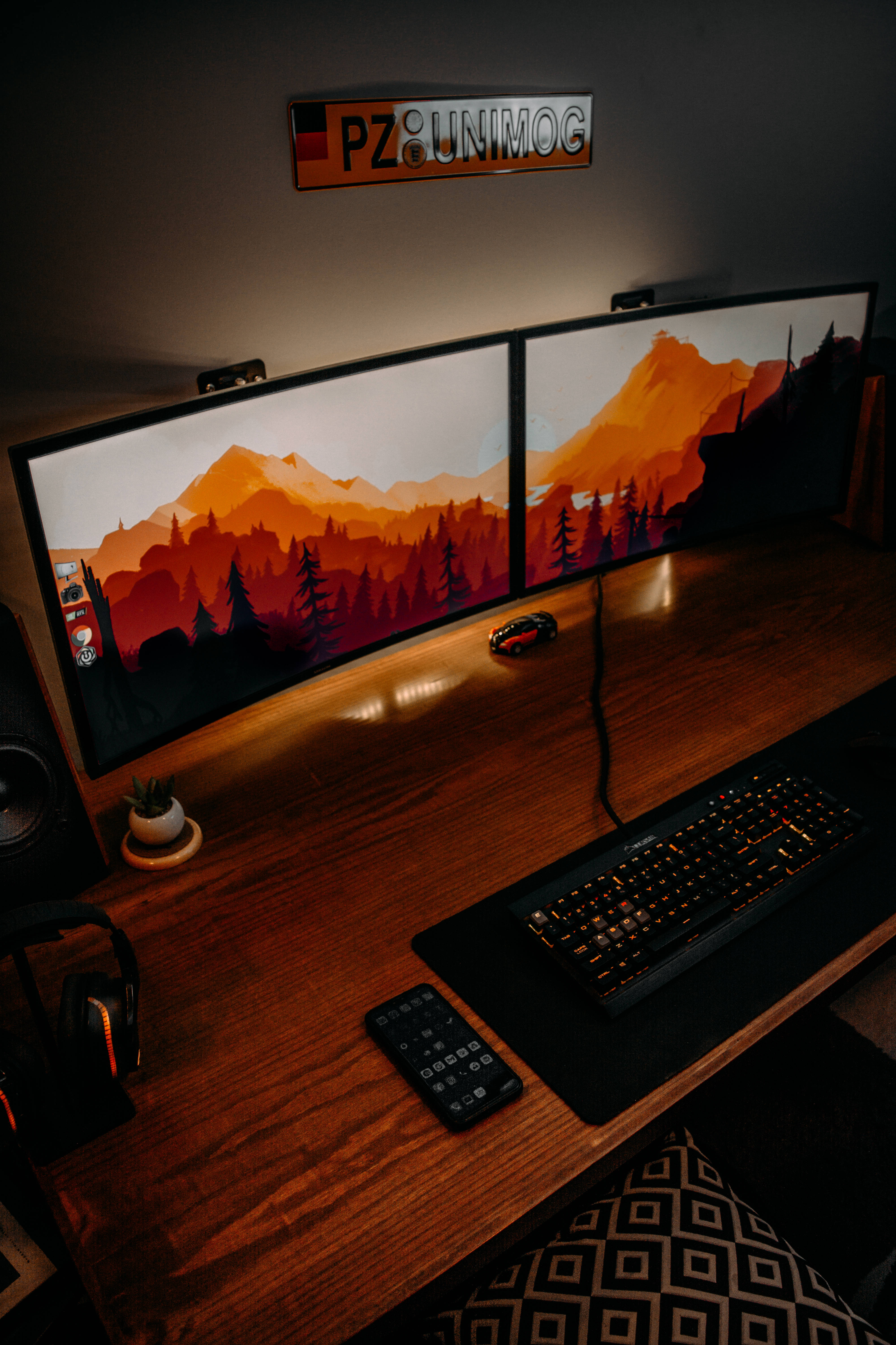 PC 遊戲 桌布 壁紙 — 一個昏暗的遊戲設置，雙顯示器顯示熾熱的山日落壁紙，木桌上配有發光的鍵盤和滑鼠。