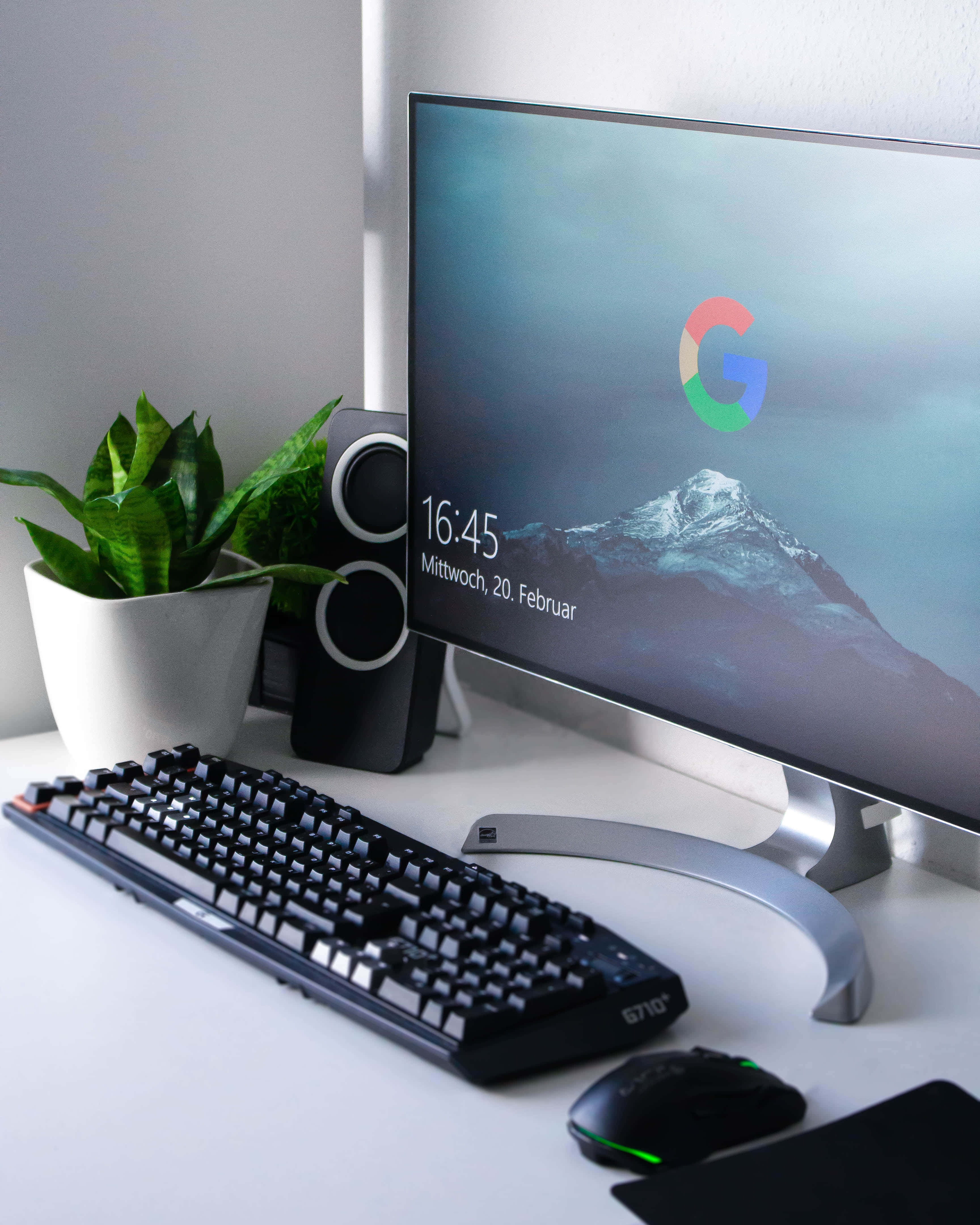 Monitor 桌布 壁紙 — 一個時尚現代的辦公桌設置，配備曲面顯示器顯示山脈壁紙、黑色鍵盤、發光滑鼠和盆栽植物，在柔和自然光下。