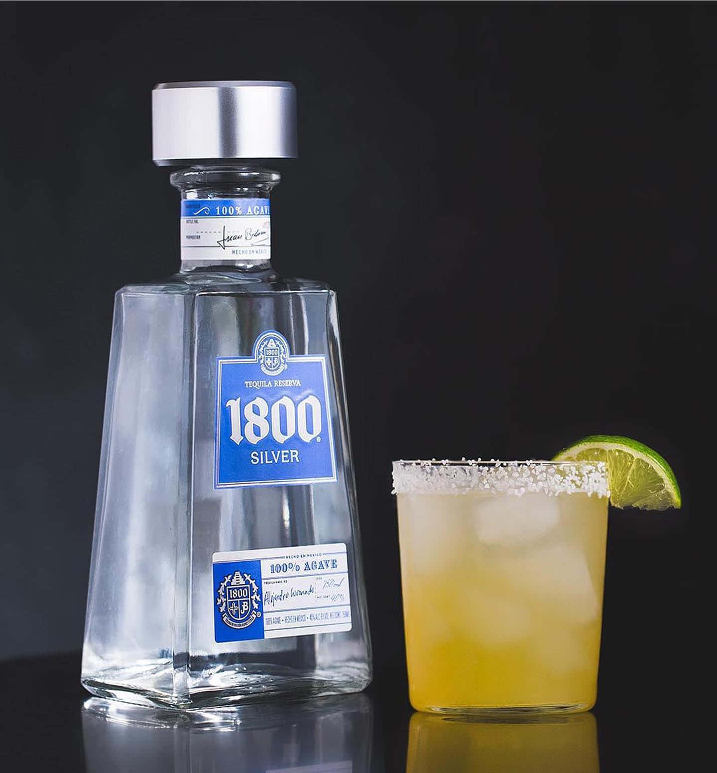 alcohol 桌布 壁紙 — 一個時尚的1800 Silver龍舌蘭酒瓶旁邊放著一杯鹽邊瑪格麗特，配以萊姆裝飾，置於深色反光表面上。