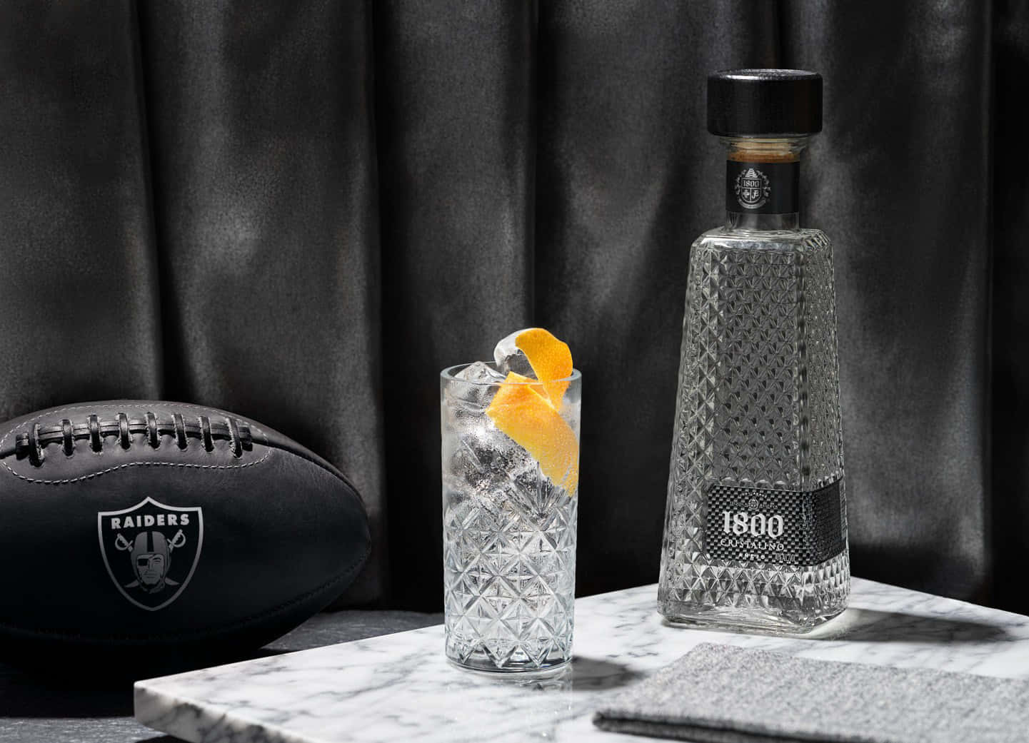 alcohol 桌布 壁紙 — 一個時尚的1800龍舌蘭酒瓶和帶有橙子裝飾的雞尾酒，旁邊是一個黑色突襲者橄欖球，放在大理石表面上。