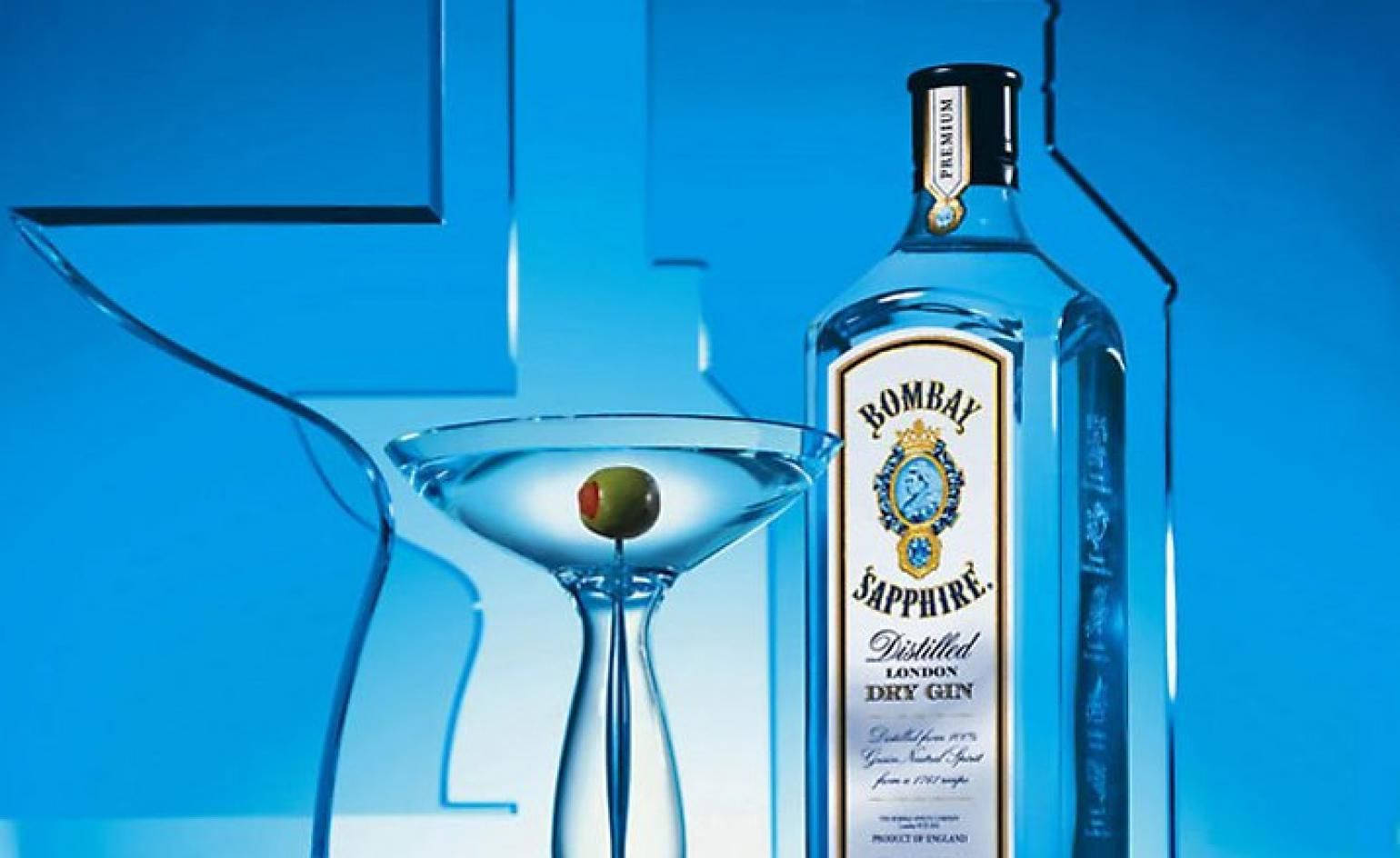 葡萄酒 桌布 壁紙 — 一個時尚的Bombay Sapphire琴酒瓶站在一個有橄欖的馬丁尼酒杯旁邊，背景是充滿活力的藍色幾何圖案。